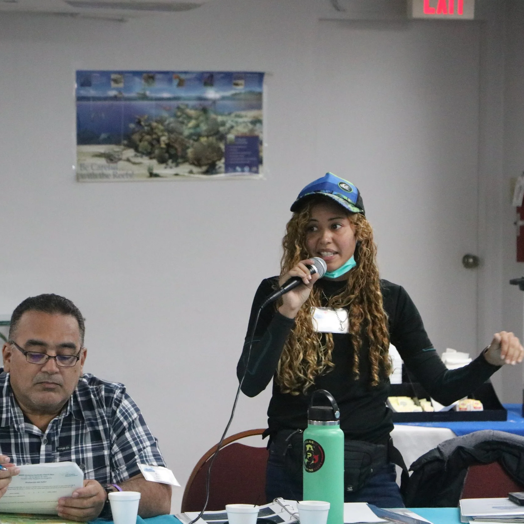 A woman with curly hair, wearing a blue cap and black shirt, is speaking into a microphone during a meeting or presentation. She has a green water bottle and a grey jacket on the table in front of her. Two men are seated at the table, one listening a