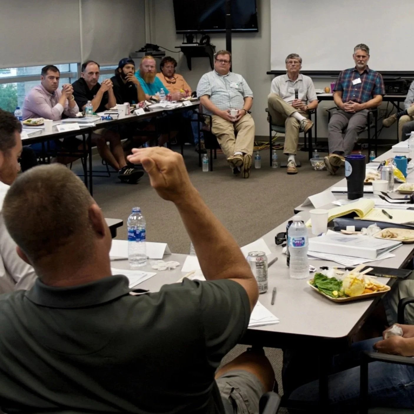 A large group of people seated in a U-shaped arrangement in a conference room, engaged in discussion or listening, with tables covered in papers, snacks, and drinks, and a large window showing an outside view.