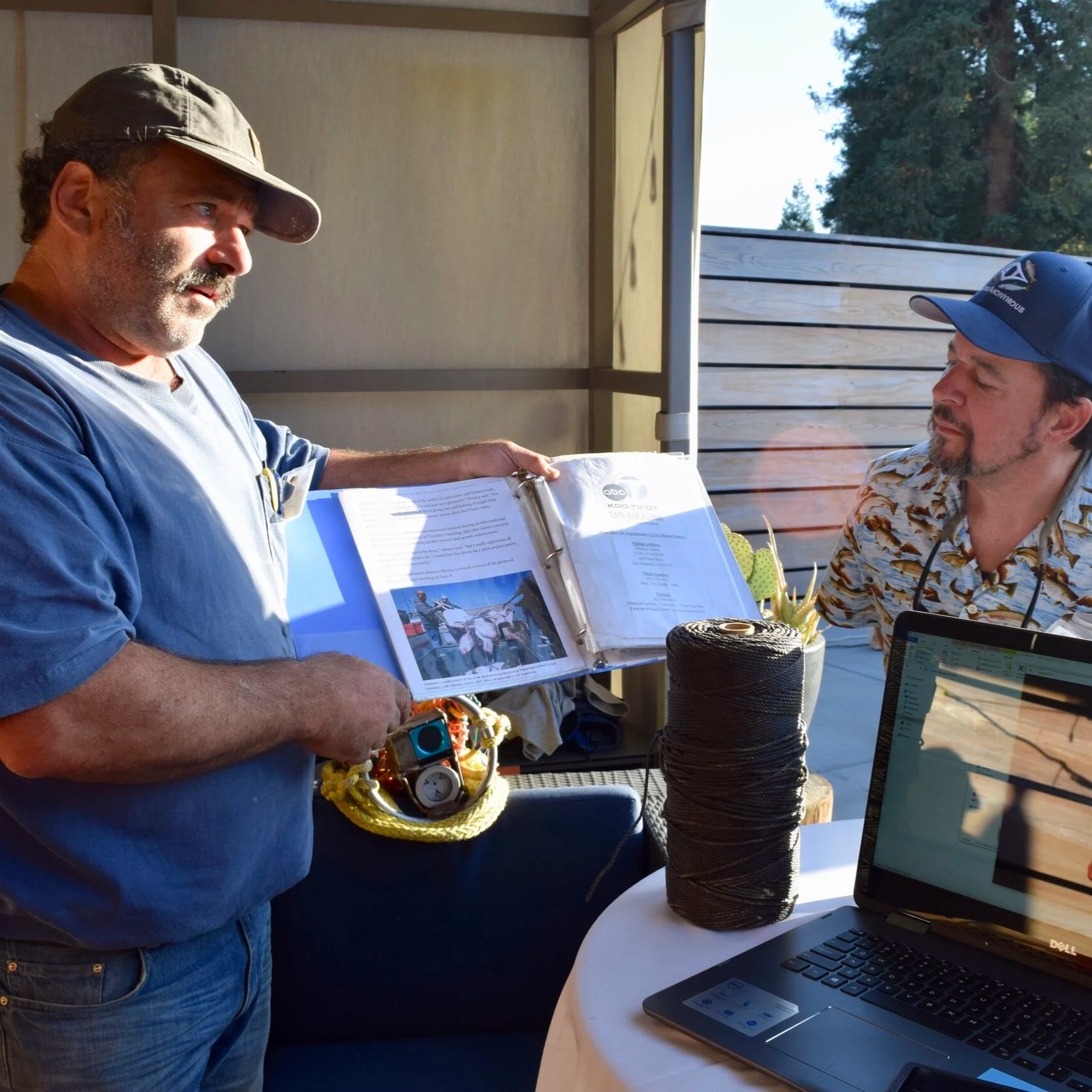 A man in a gray baseball cap and blue shirt shows a photo album to two people seated at a table, with a laptop and spool of black thread on the table; they are outdoors on a porch with string lights and trees in the background.