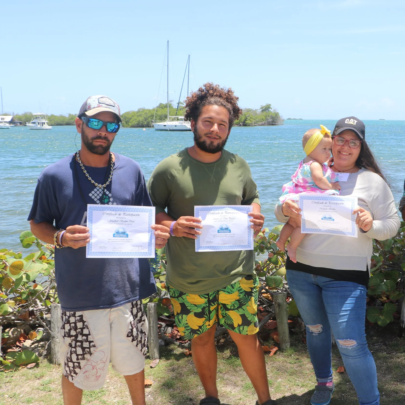 Group of five people standing outdoors near the water, holding certificates, with sailboats in the background.