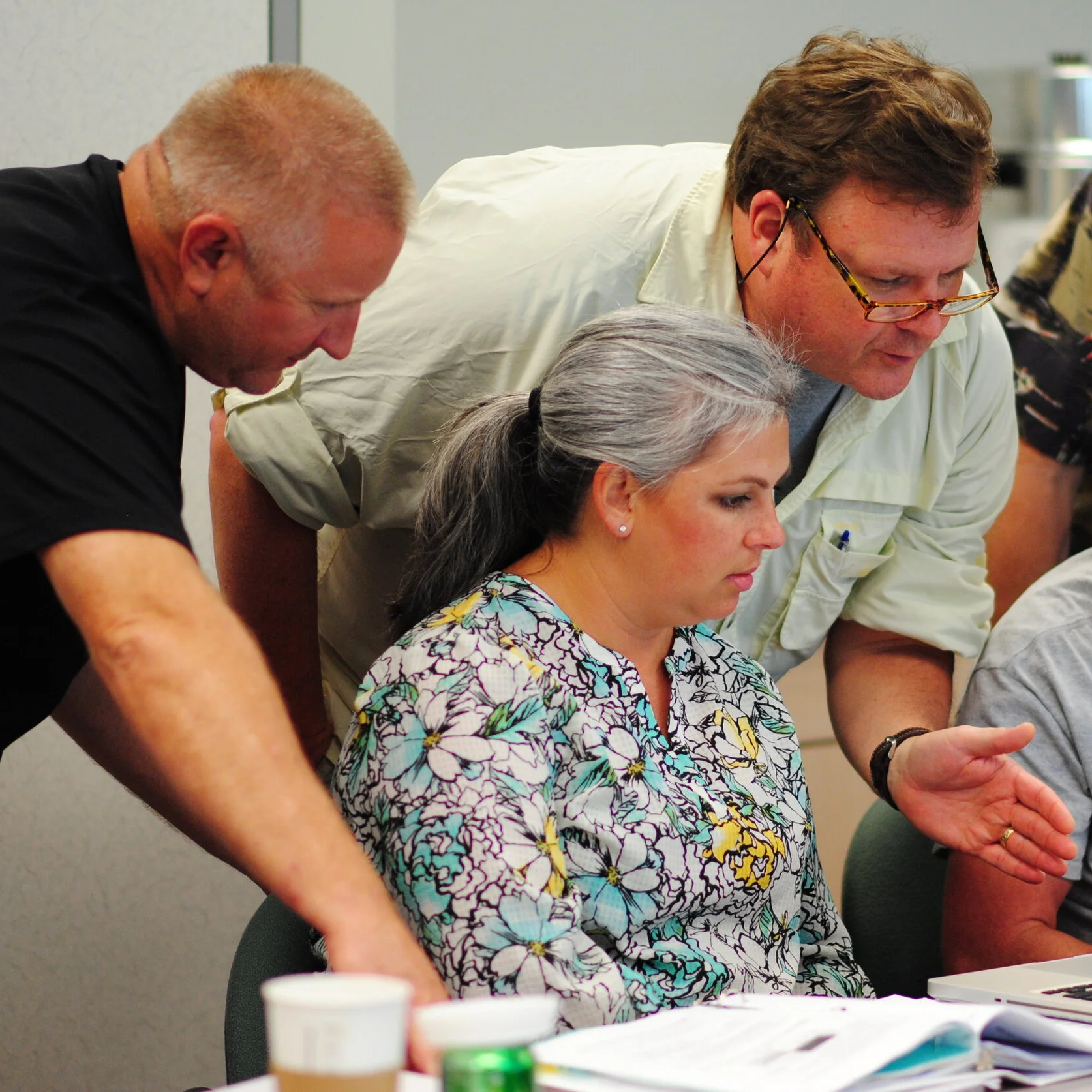 Five people gathered around a laptop, examining the screen together, with papers and coffee cups on the table.