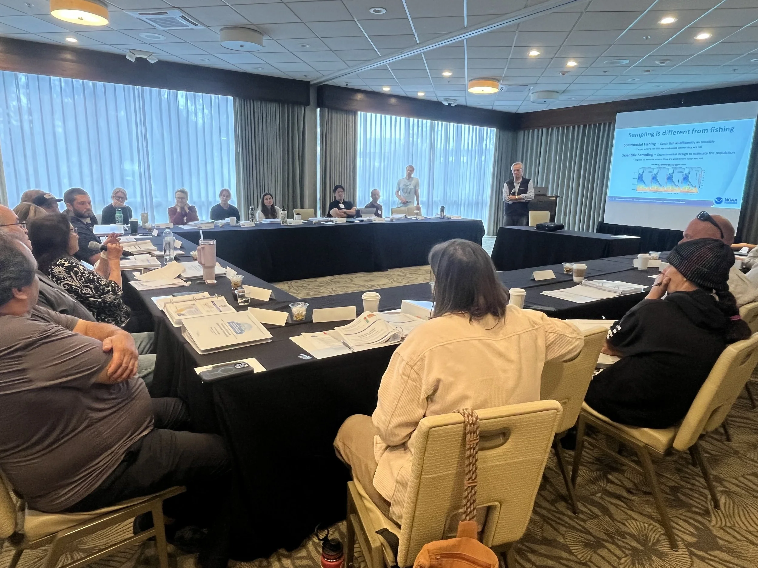 Conference attendees are sitting around a large table listening to a presentation that's projected on a screen