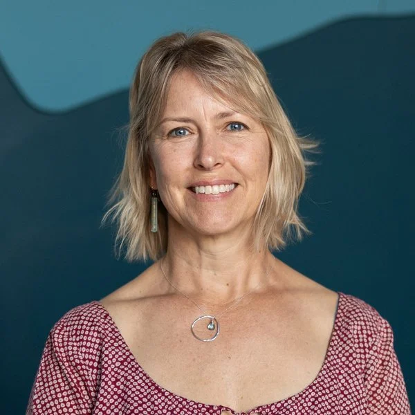 A woman with short blonde hair, wearing a black headband with sunglasses on top, a red jacket, and a black neck warmer, smiling in front of a lake with snowy mountains in the background.