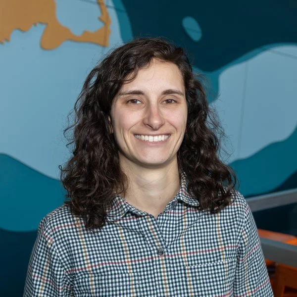 A woman with curly brown hair smiling outdoors in front of stacked wire lobster traps, with a blue sky, houses, and trees in the background.