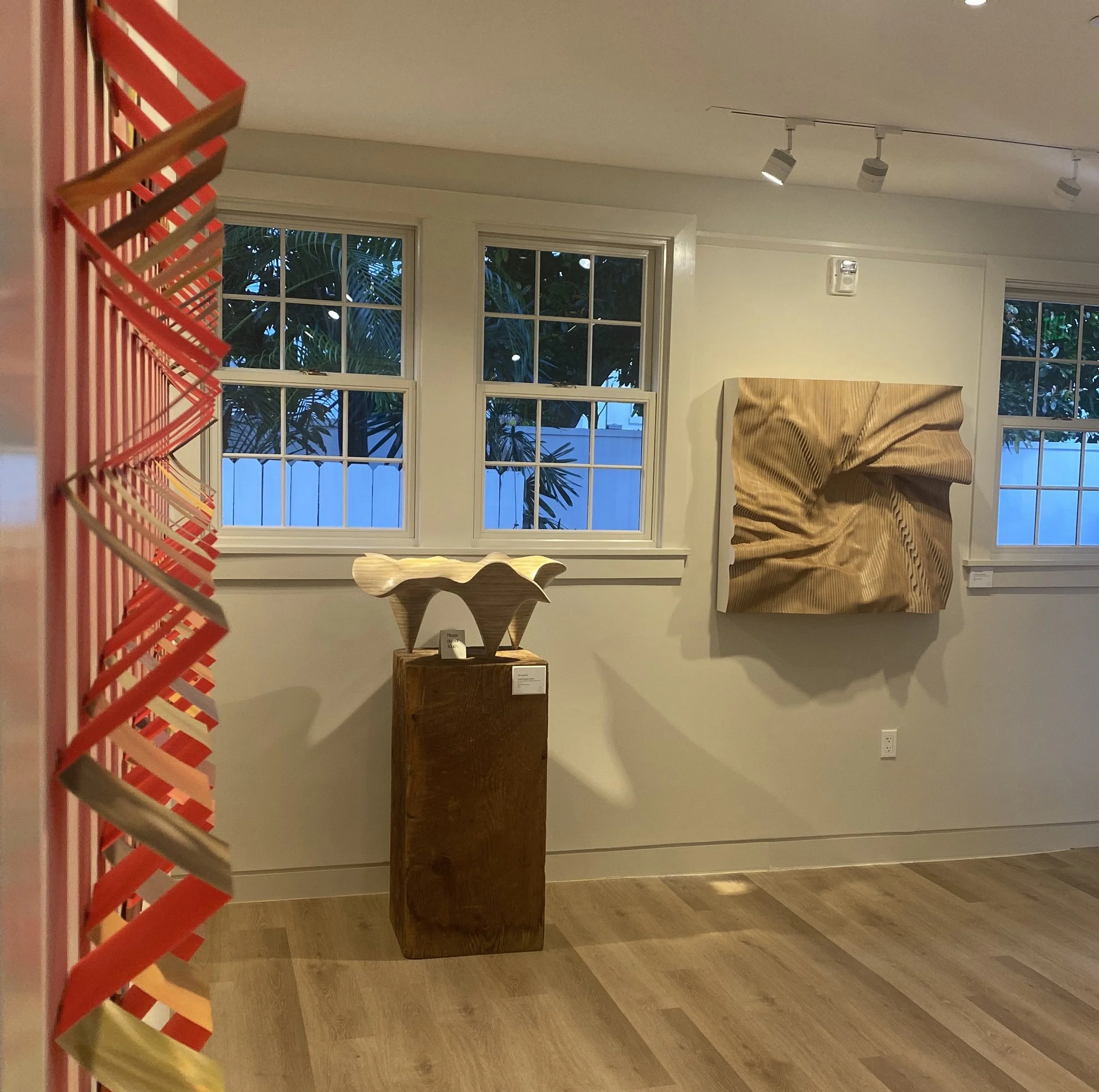 Art gallery at the Ann Norton Sculpture Gardens, with wooden sculpture on wood pedestal, wood sculpture on the wall, paper sculpture from side, and windows showing trees outside.