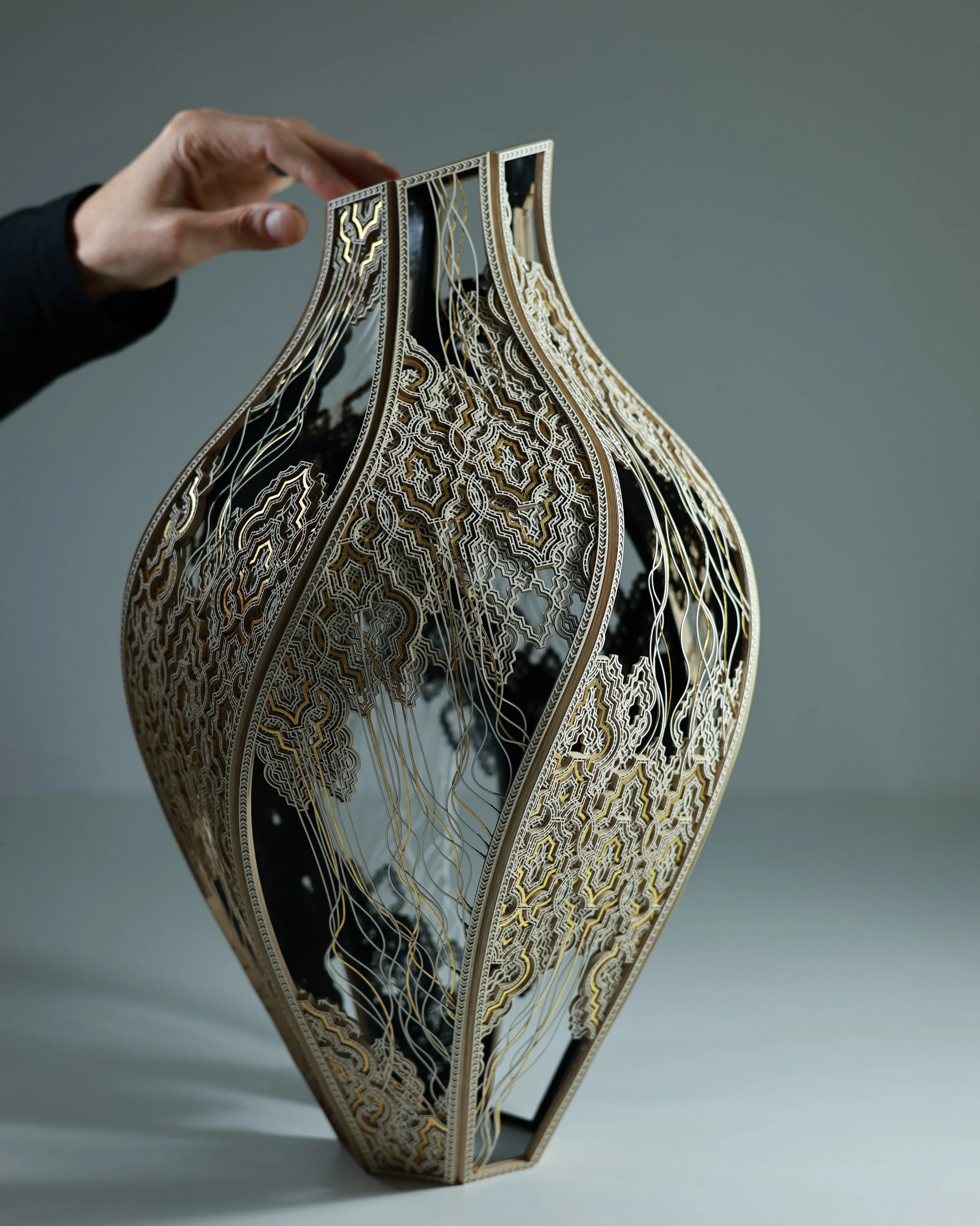 A full vertical image of Julia Ibbini’s 'Kŷma | Vessel'. The work is comprised of paper, ink, cotton, and gold leaf. The hand in the frame illustrates the scale of the work.
