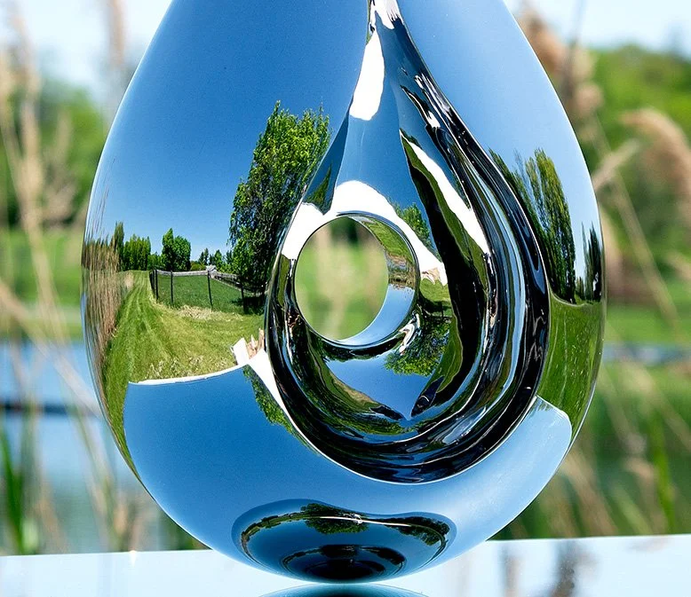 A detail from Tarik Currimbhoy’s kinetic stainless steel sculpture, 'Paisley', shown on top of a mirrored base against a natural backdrop.