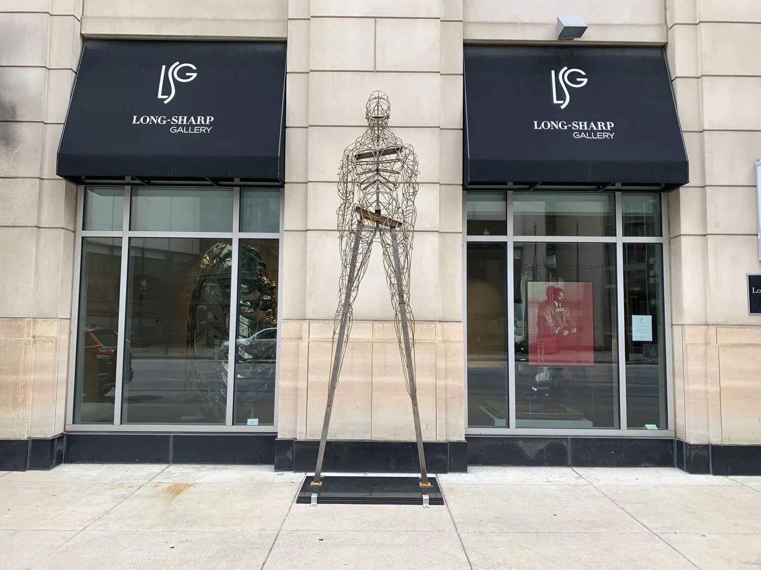 A large metal sculpture of a human figure standing outside the Long-Sharp Gallery, between two glass windows with black awnings.