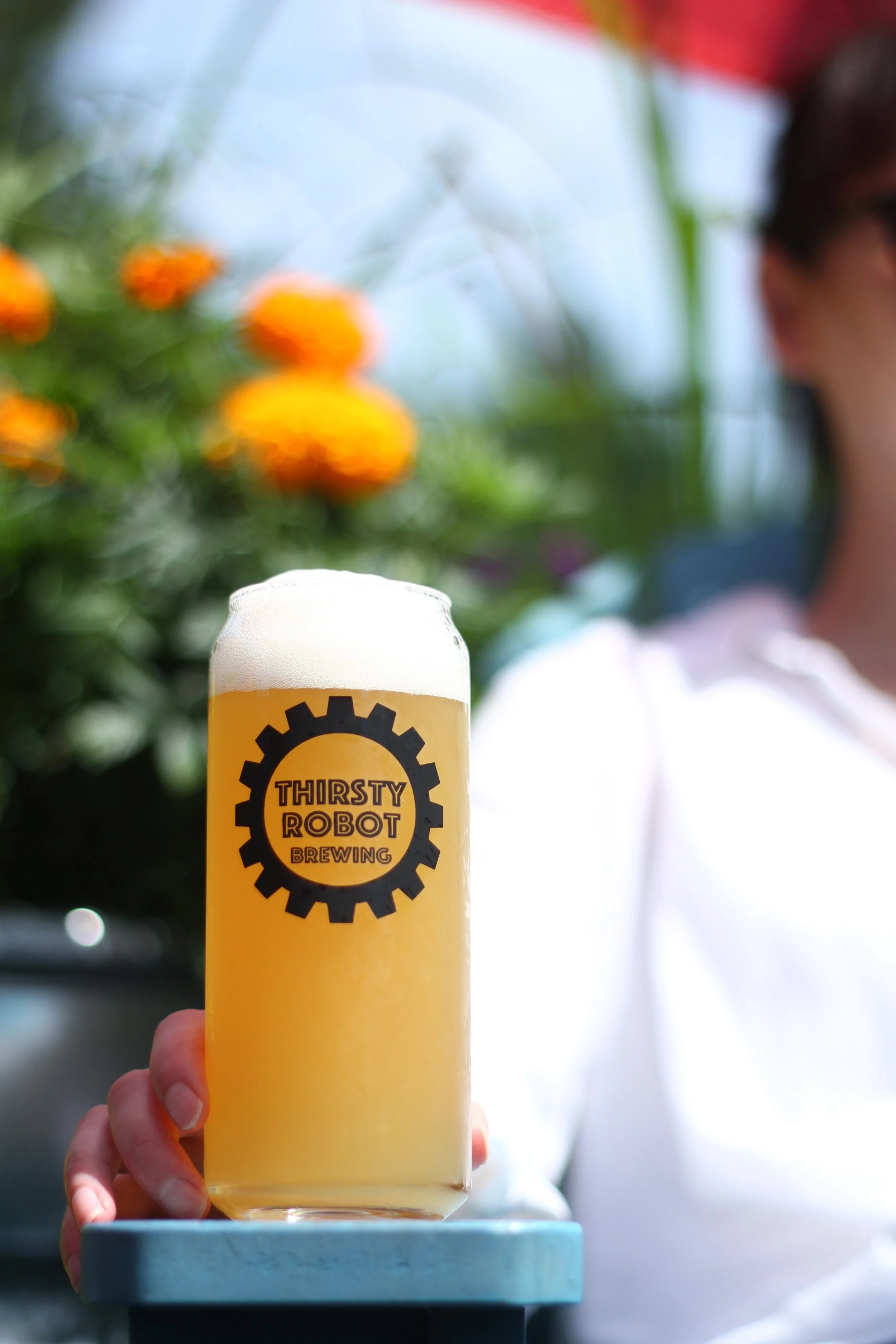 A glass of beer with a foam head, branded with the Thirsty Robot Brewing logo, being held by a person outdoors with blurred plants and a woman in the background.