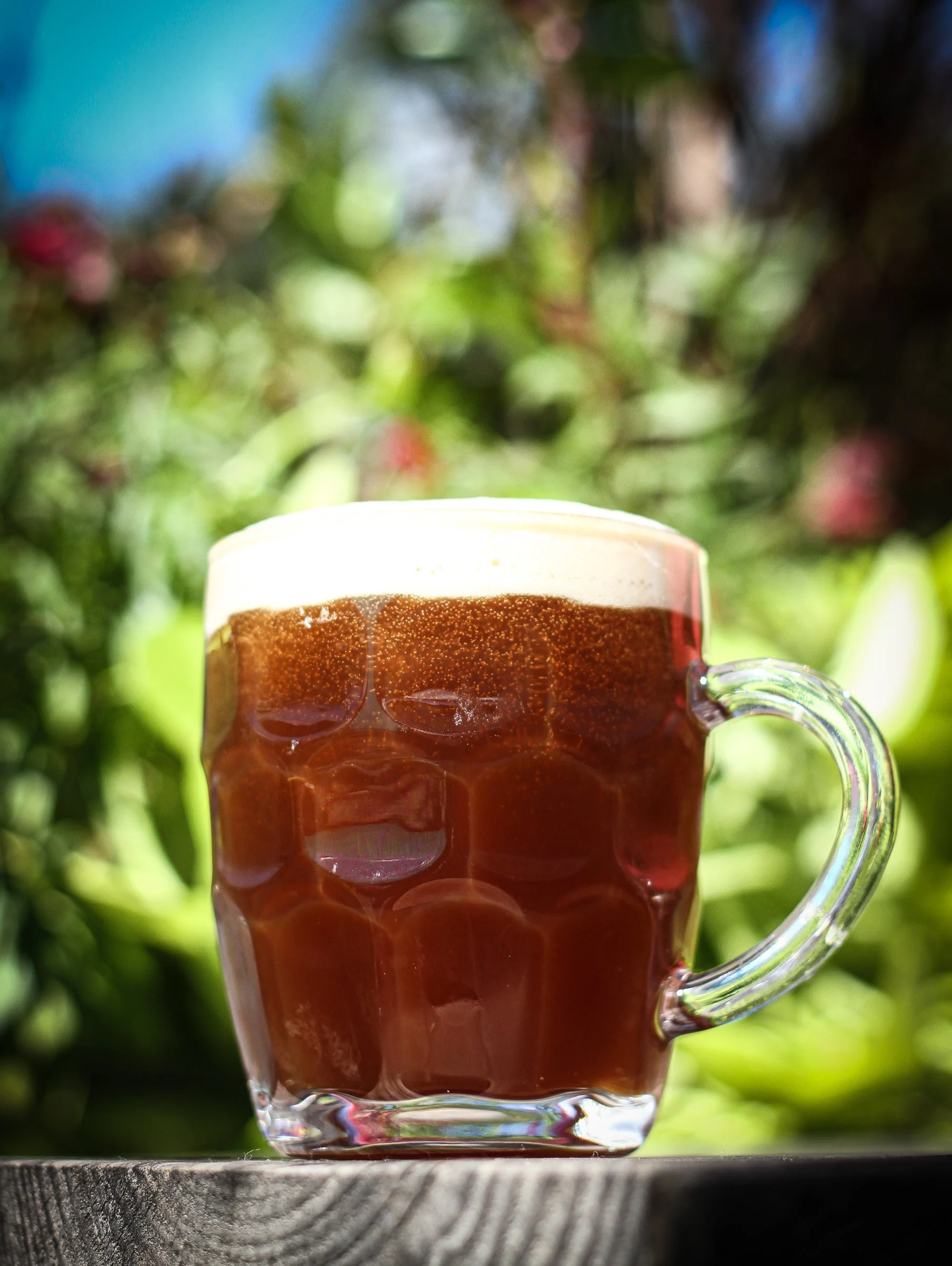 A glass mug filled with dark beer with a foamy head, placed on a wooden surface outdoors with blurred greenery in the background.