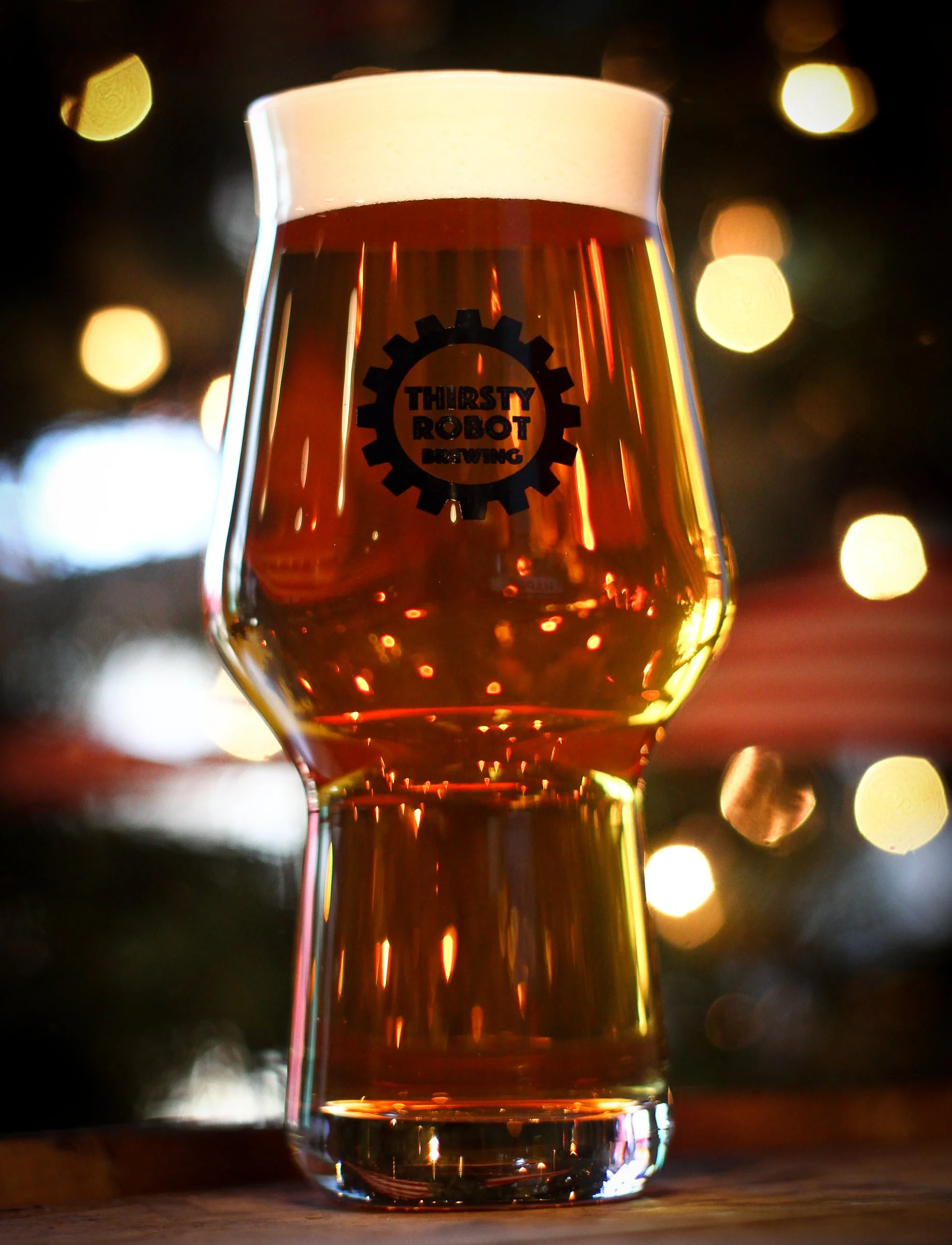 A glass of amber-colored beer with a foamy head, featuring a logo that reads "Thirsty Robot Brewing" on the front, placed on a wooden surface with a blurred background of warm bokeh lights.