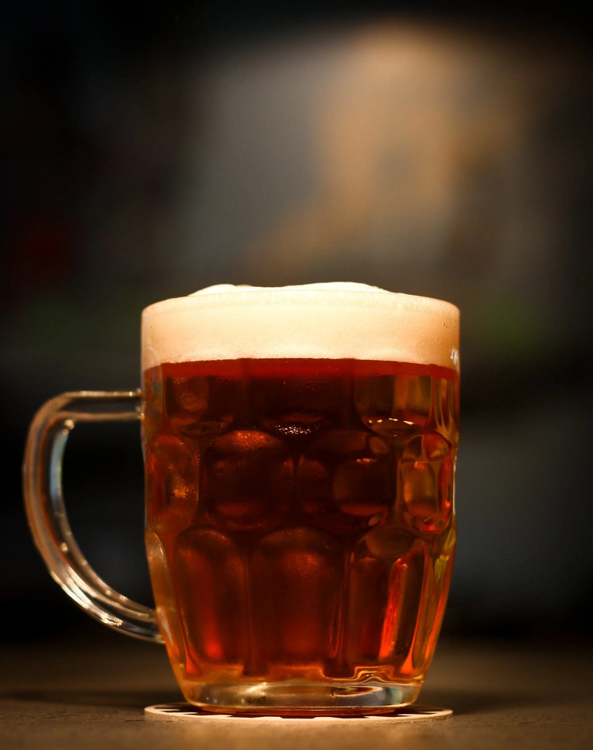 A glass mug filled with amber-colored beer topped with a foamy head, placed on a dark surface with a blurred background.