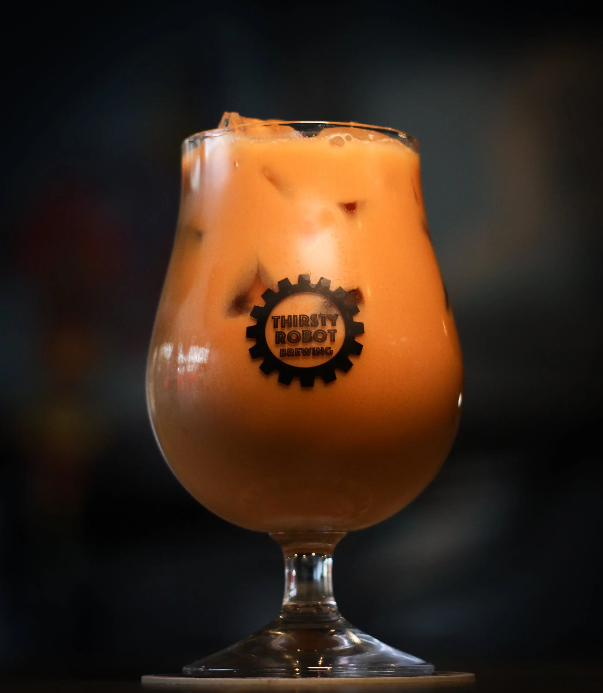 A glass with an orange-colored beverage filled with ice, labeled 'Thirsty Robot Brewing', against a blurred dark background.