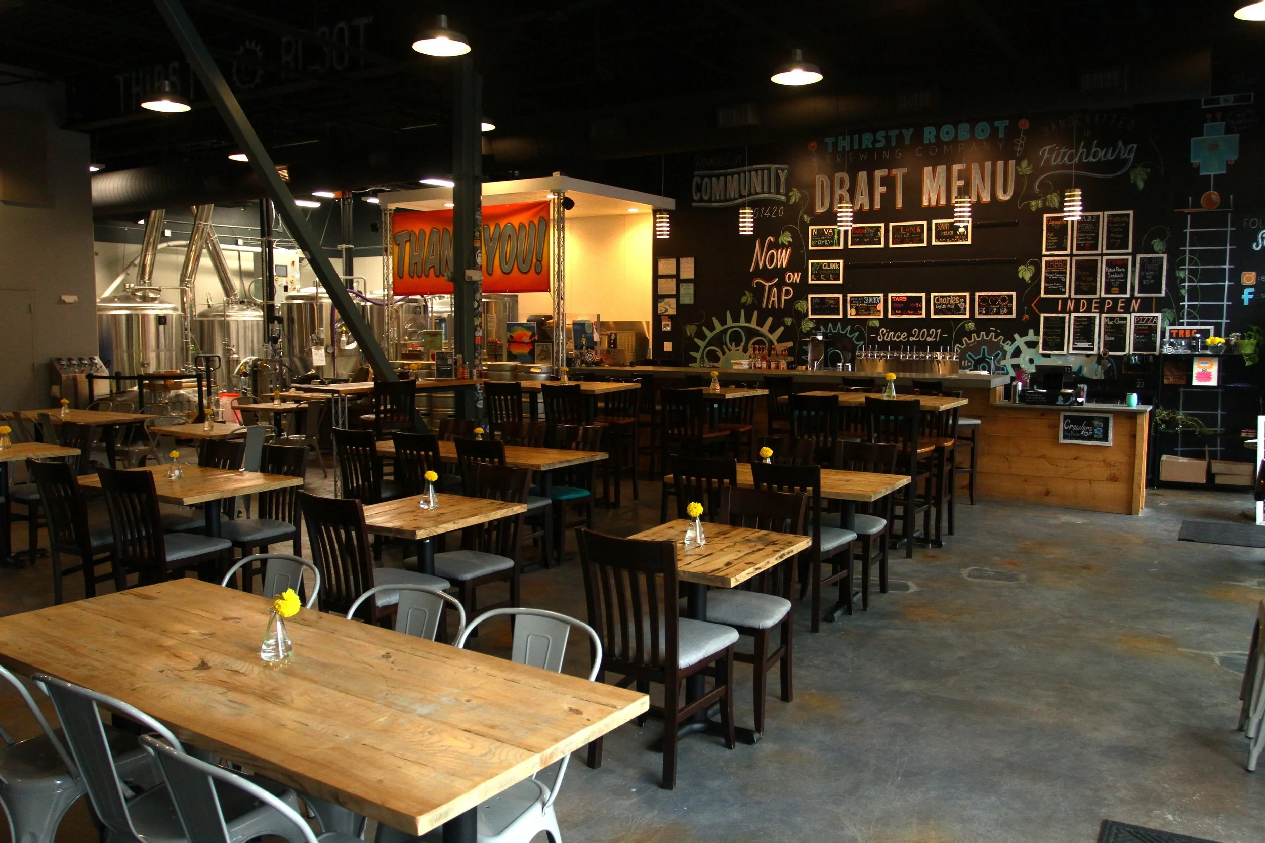 Inside view of a brewery or taproom with wooden tables, metal and wooden chairs, a chalkboard menu on the wall, and brewing tanks in the background, decorated with small vases of yellow flowers on each table.