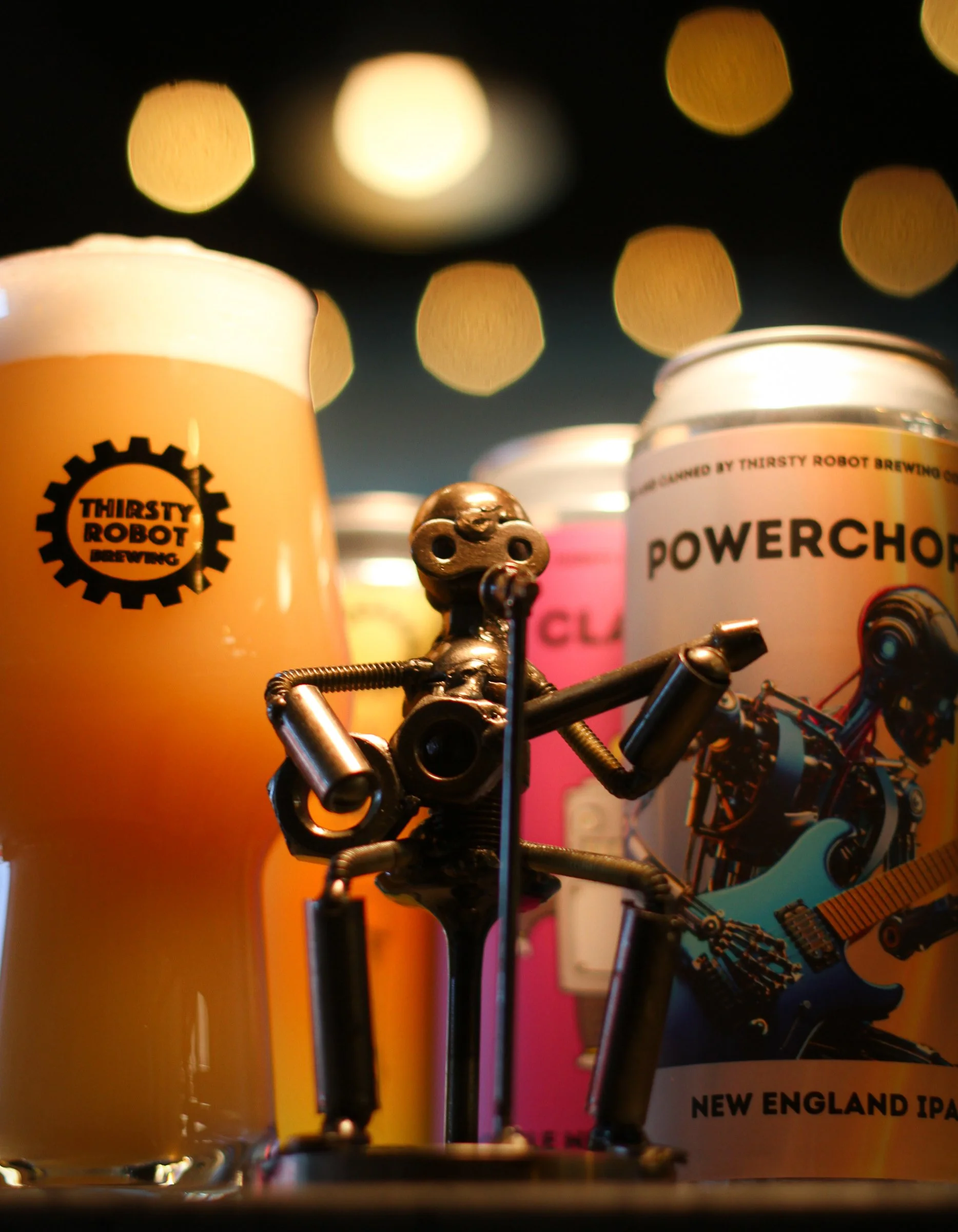 Close-up of a small robot figurine with a beer glass and a beer can in the background, featuring glowing bokeh lights in the dark.