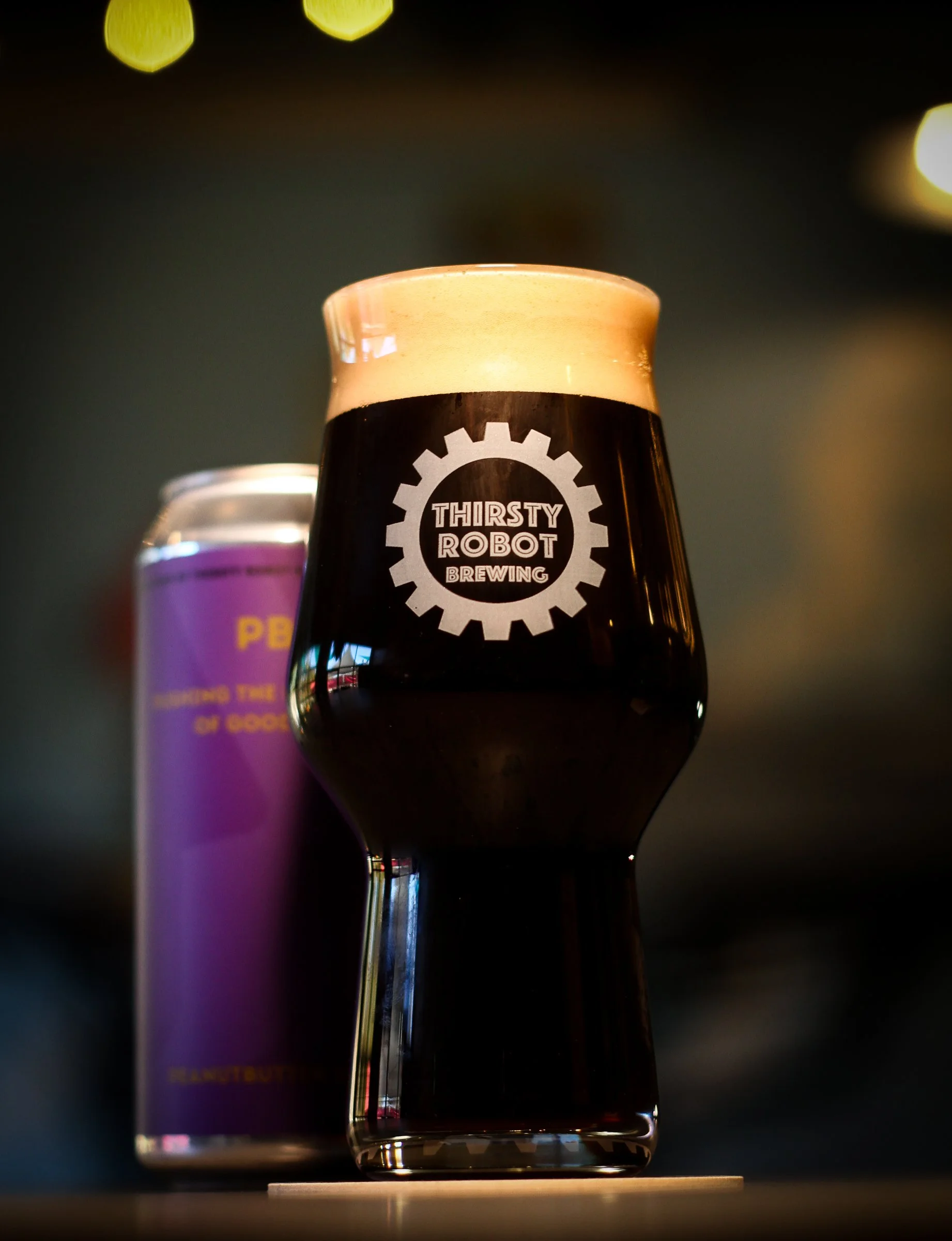 A dark beer in a branded glass with a foamy head, labeled 'Thirsty Robot Brewing.' In the background, there is a can with purple and yellow colors.