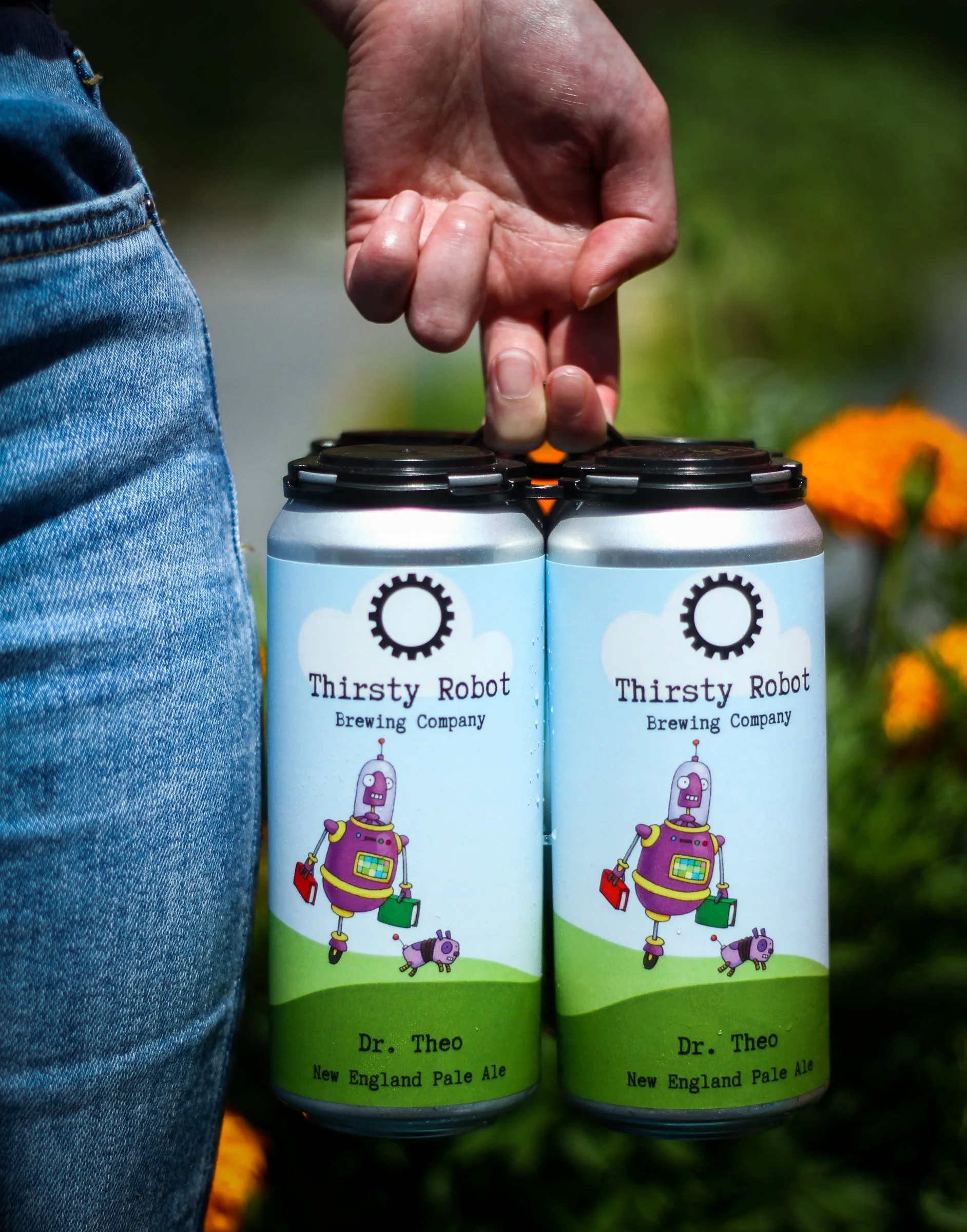 A person holding two cans of Thirsty Robot Brewing Company beer, labeled 'Dr. Theo' New England Pale Ale, with colorful robot illustration, outdoors with blurred green background and orange flowers.