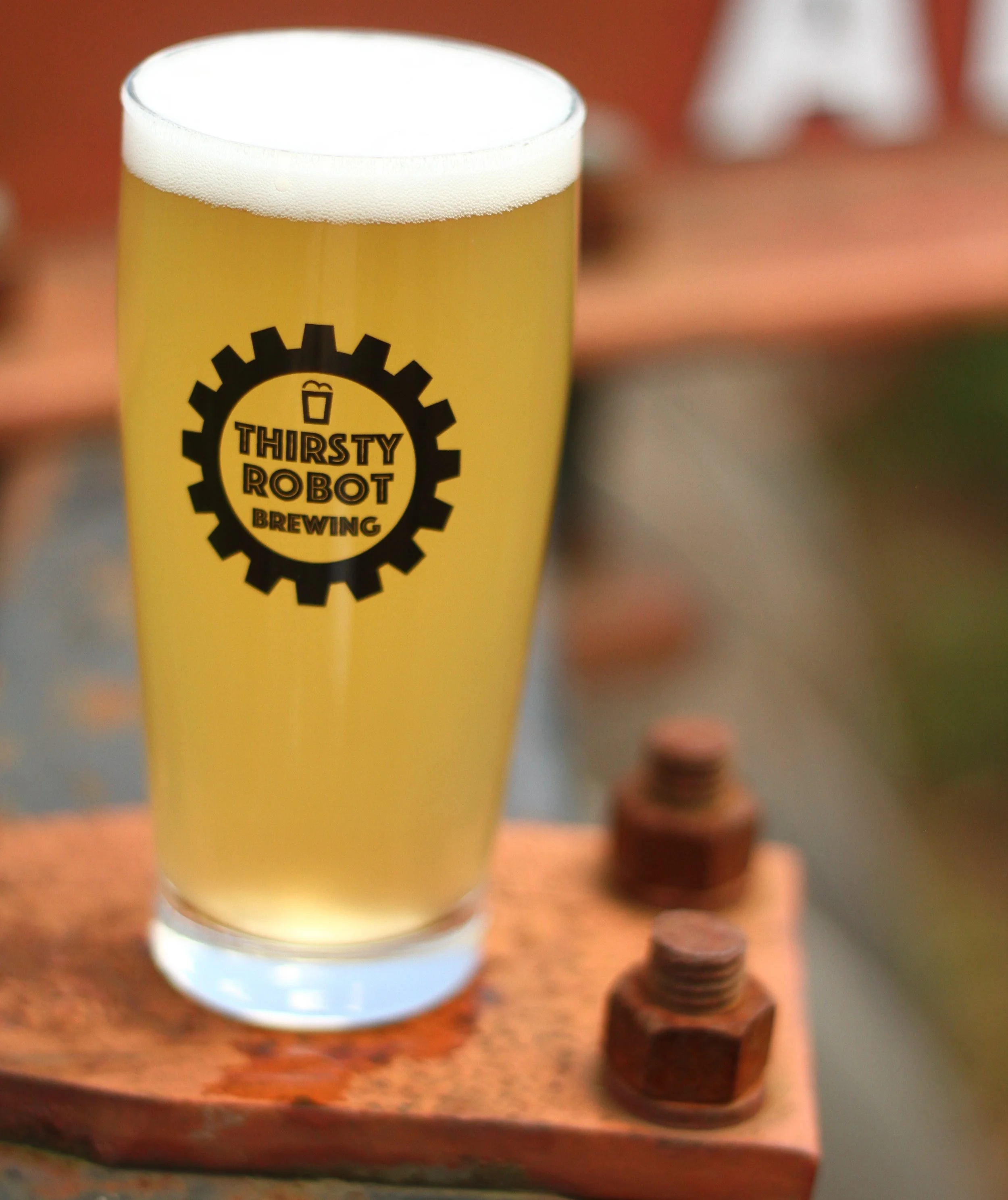 A glass of beer with a logo of Thirsty Robot Brewing on it, placed on a metal surface with bolts.