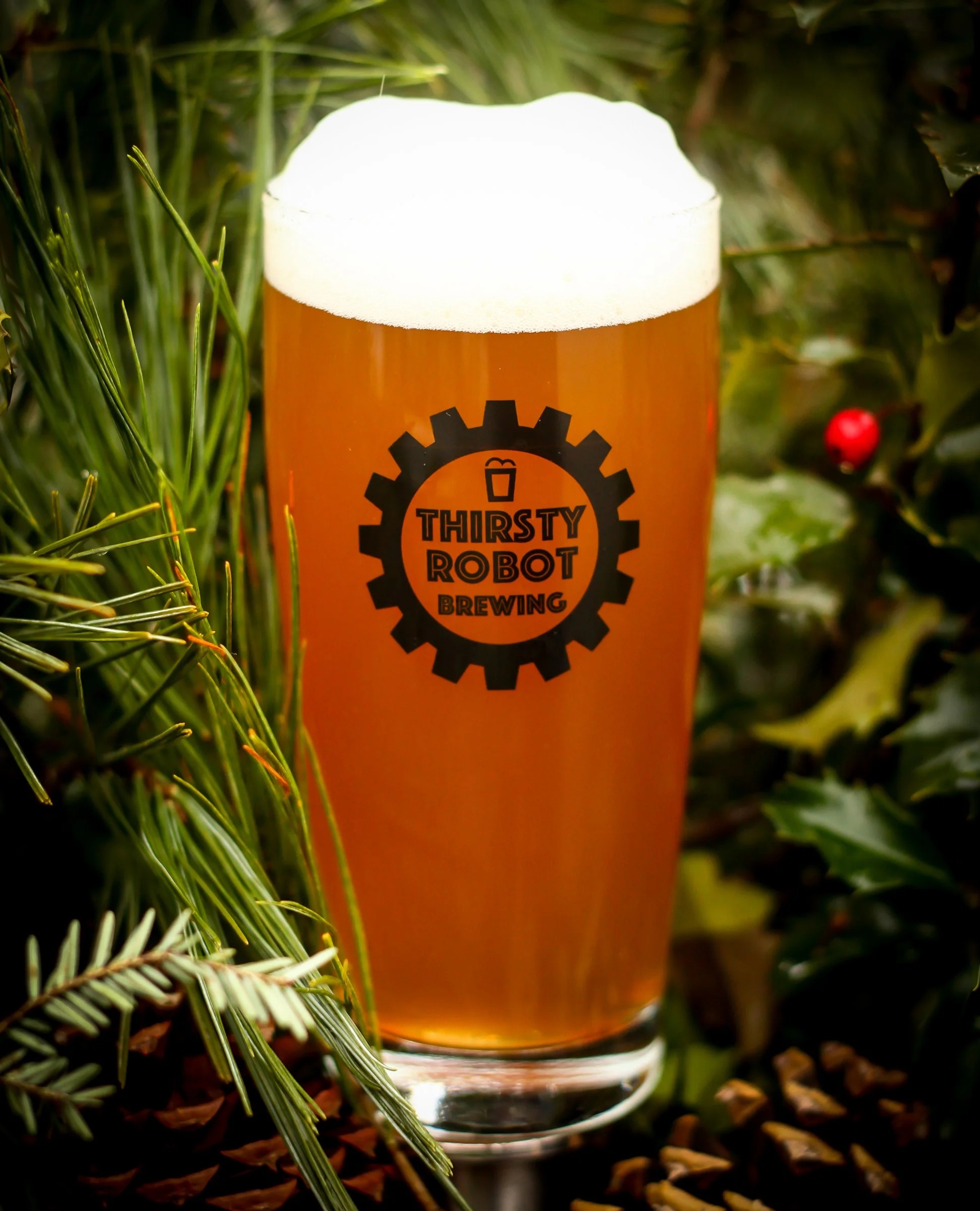 A glass of beer with a foamy head featuring a 'Thirsty Robot Brewing' logo, surrounded by greenery and holiday foliage.