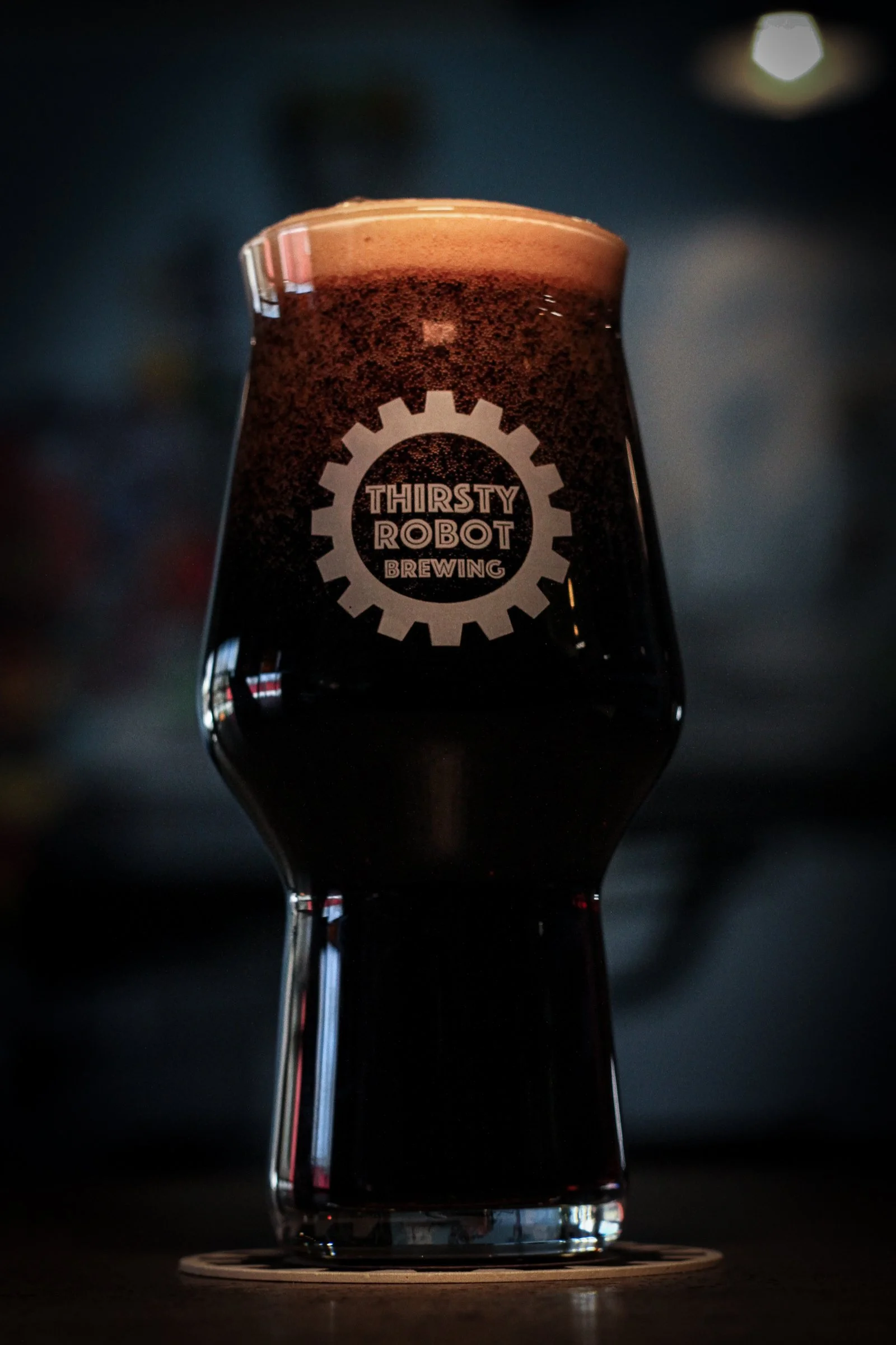 A dark beer in a tulip-shaped glass with 'Thirsty Robot Brewing' logo on it, placed on a wooden coaster, against a blurred background.