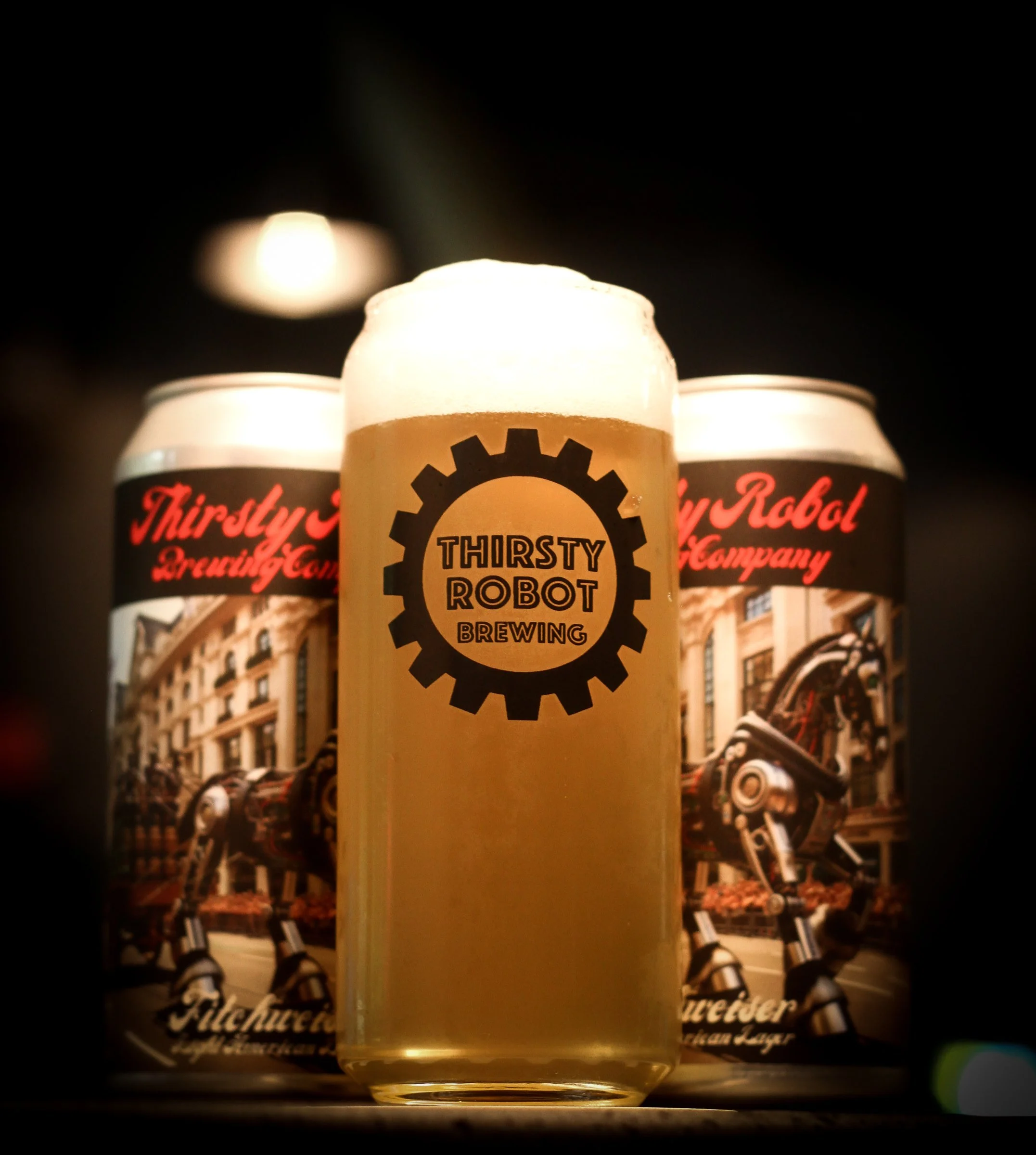 A glass of beer with foam on top, labeled Thirsty Robot Brewing, flanked by two cans with Thirsty Robot Brewing artwork, showing a robot on a city street.