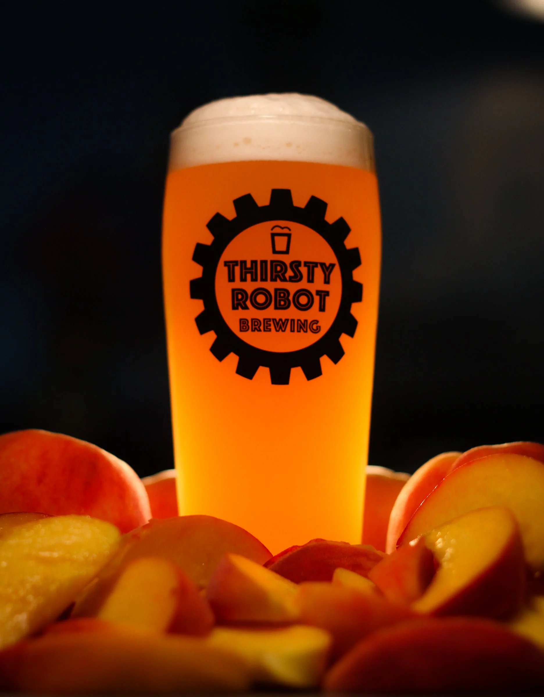 A tall glass of beer with a frothy head, featuring the Thirsty Robot Brewing logo, surrounded by sliced peaches against a dark background.