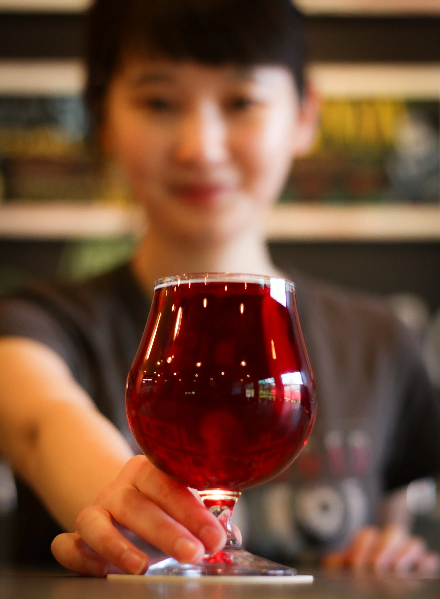 A person holding a glass of red wine, with a blurred background of shelves with various items.