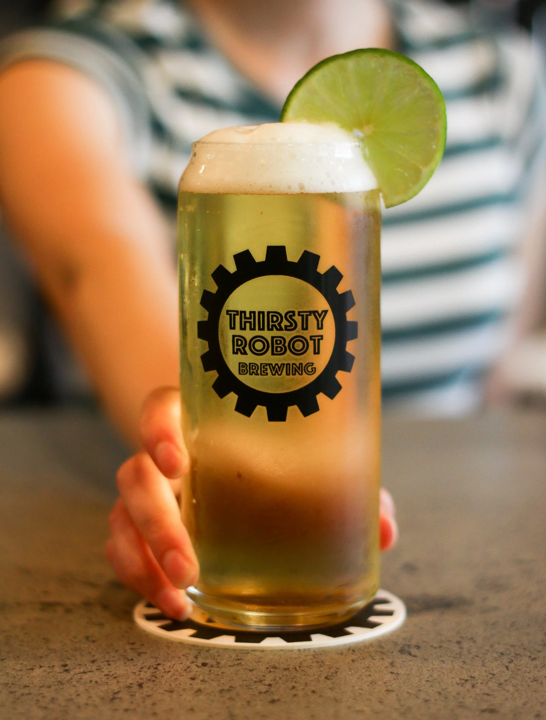 A glass of beer with foam and a lime wedge garnish, featuring the Thirsty Robot Brewing logo, held by a person in a striped shirt.