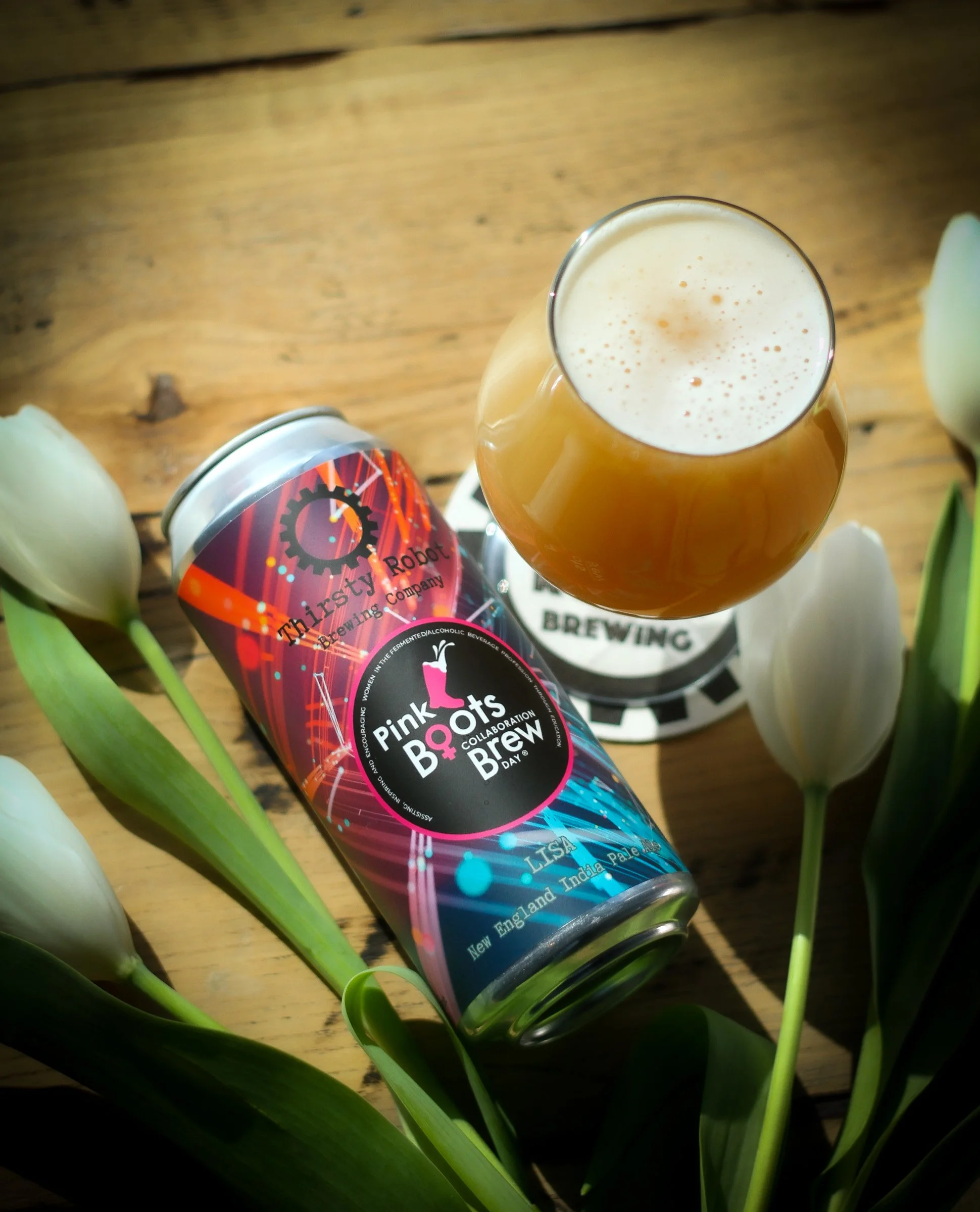 A can of Pink Boots Collaboration Brew beer labeled 'Lisa,' a glass of beer with foam, and white tulips arranged on a wooden surface.