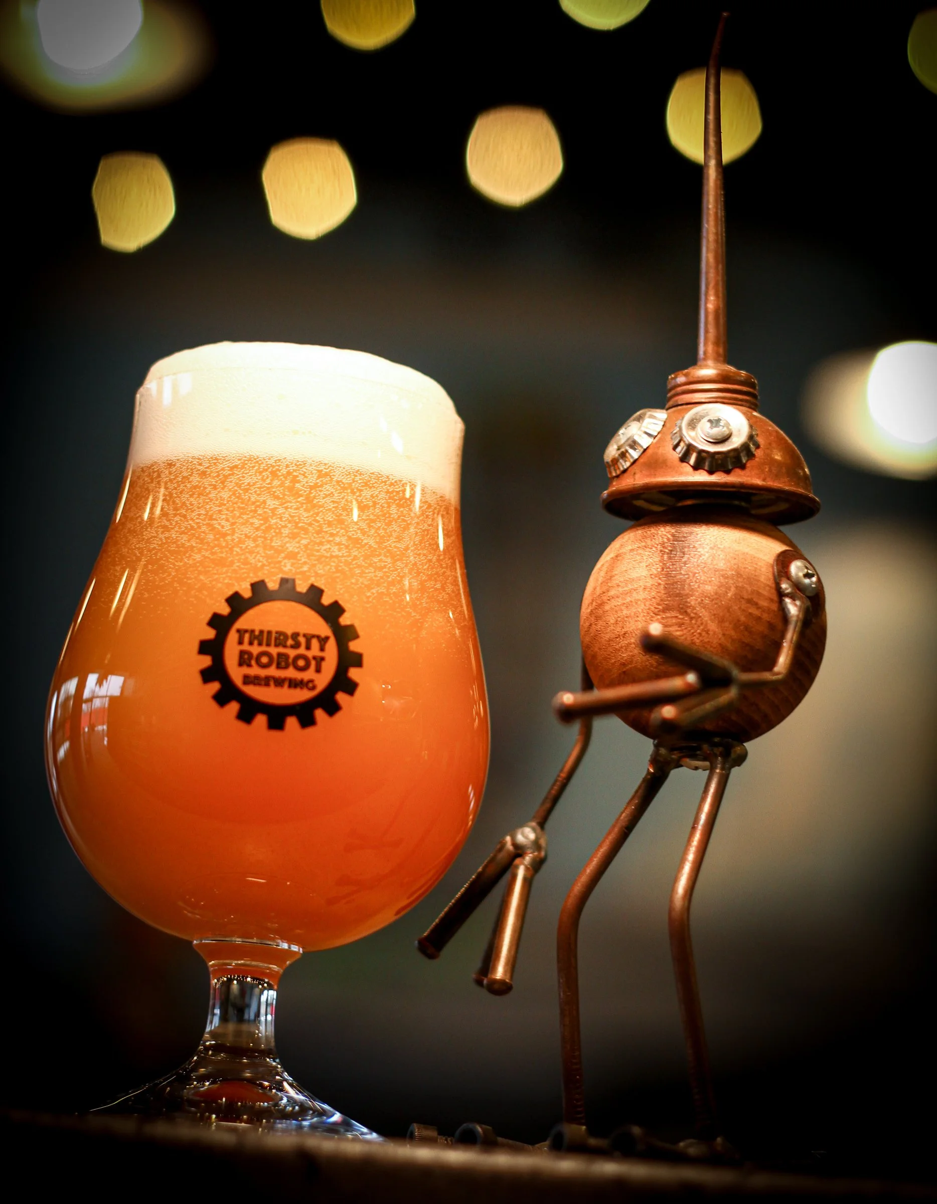 A glass of hazy, orange beer with a foam head, featuring a 'Thirsty Robot' logo, is placed next to a whimsical robot sculpture made of wood and metal with a long antenna against a blurred background with bokeh lights.