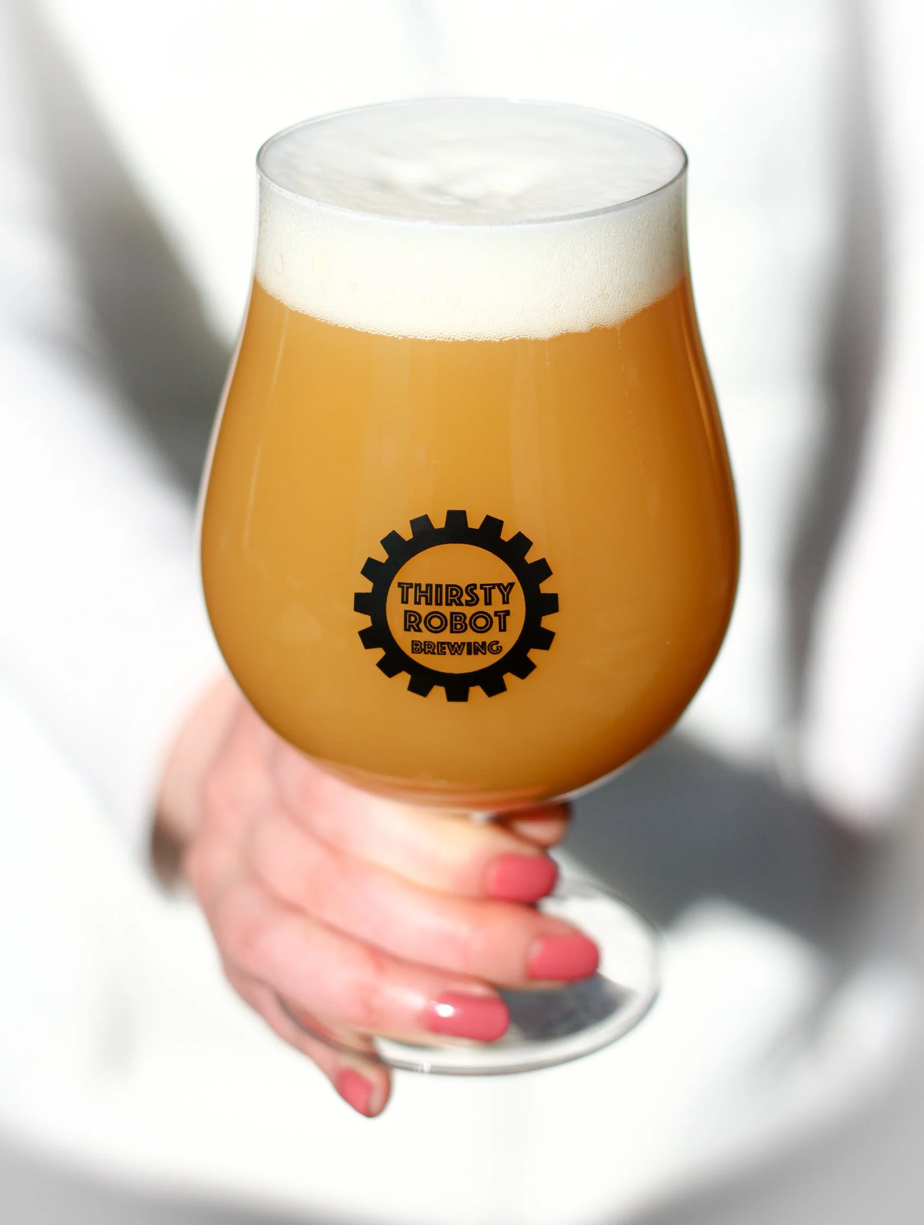 A hand holding a tulip-shaped beer glass filled with hazy, pale yellow beer, topped with a foamy white head, with the logo 'Thirsty Robot Brewing' on the glass.
