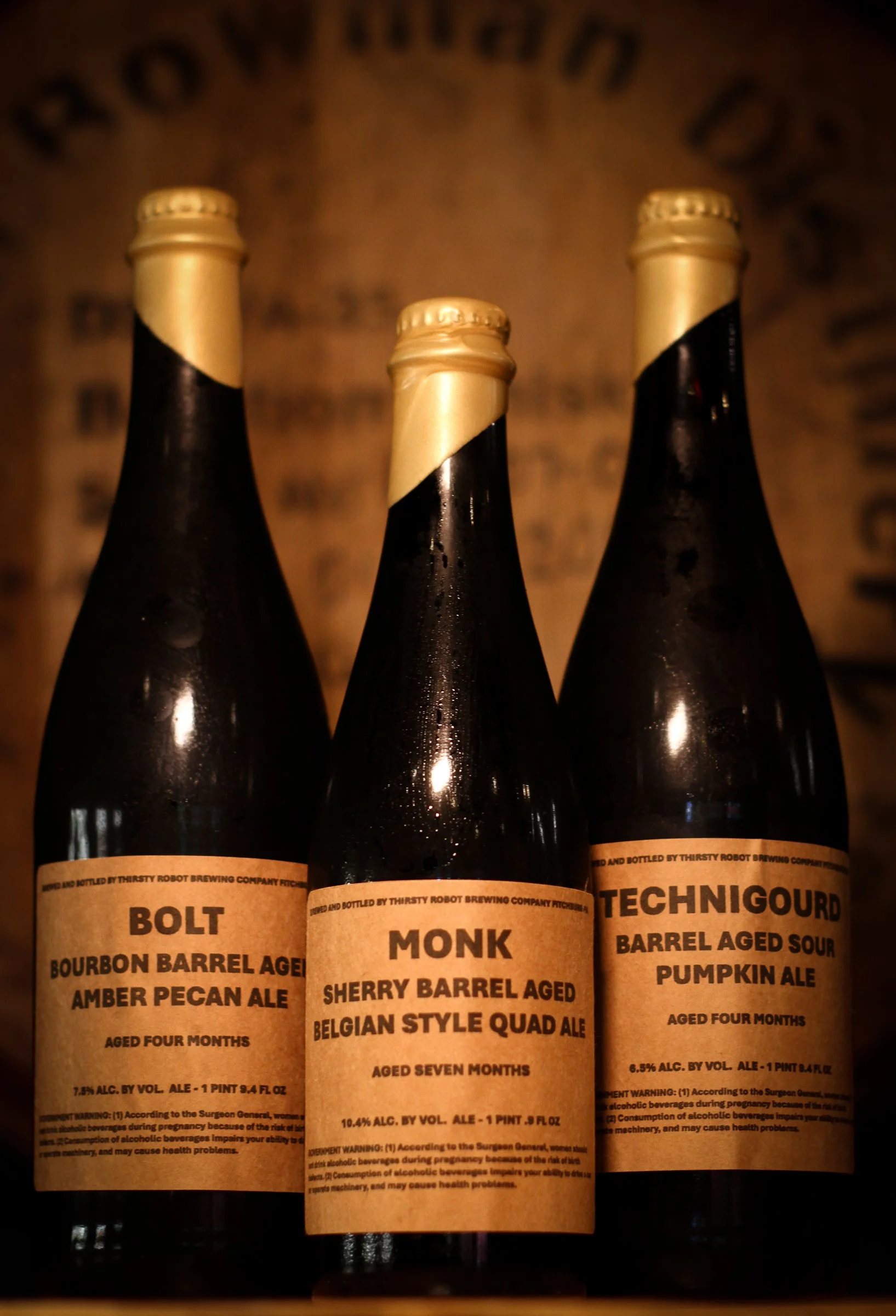 Three dark bottles of craft beer with tan labels, showing names and descriptions of different aged barrel sour beers, against a blurred background.