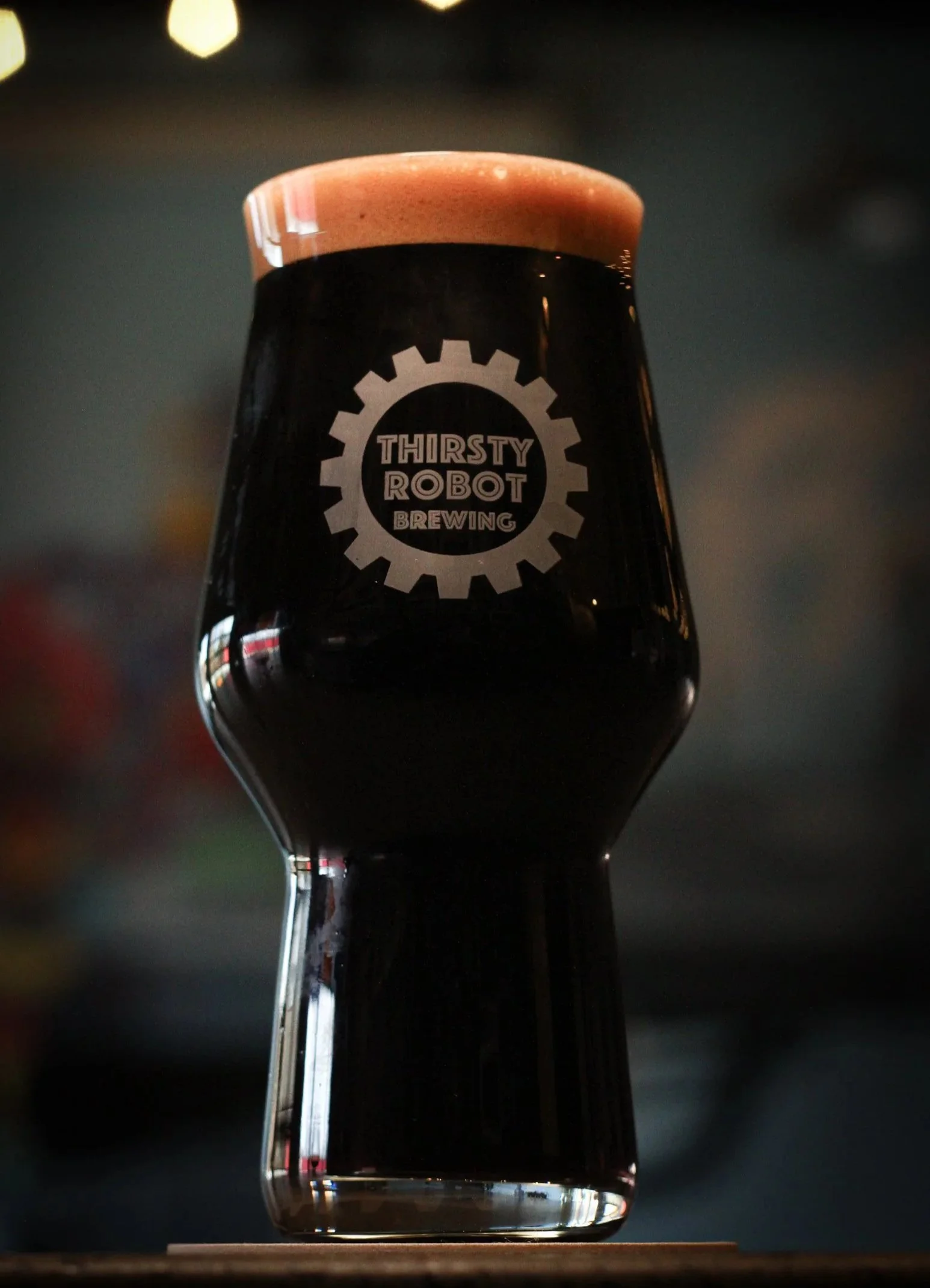 A dark beer in a glass with a foam head, labeled 'Thirsty Robot Brewing', against a blurred background.