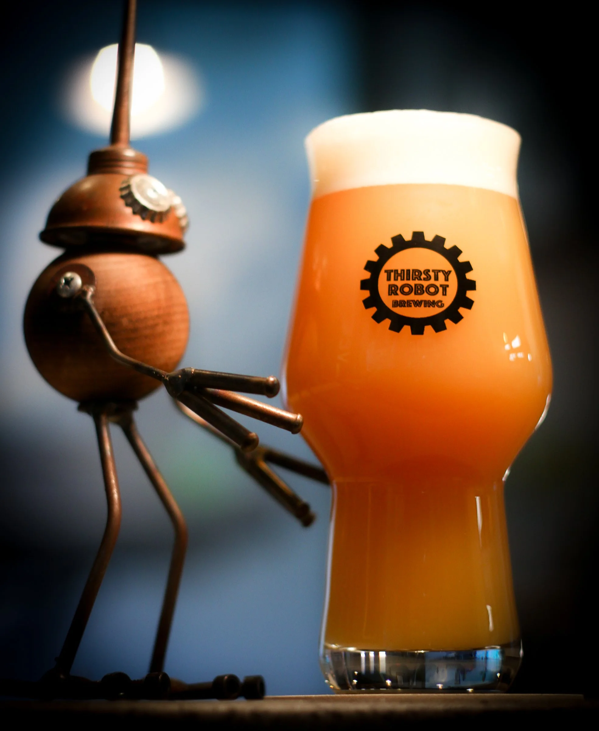 A robot sculpture holding a beer glass with the Thirsty Robot Brewing logo, filled with hazy beer topped with foam, on a dark background.
