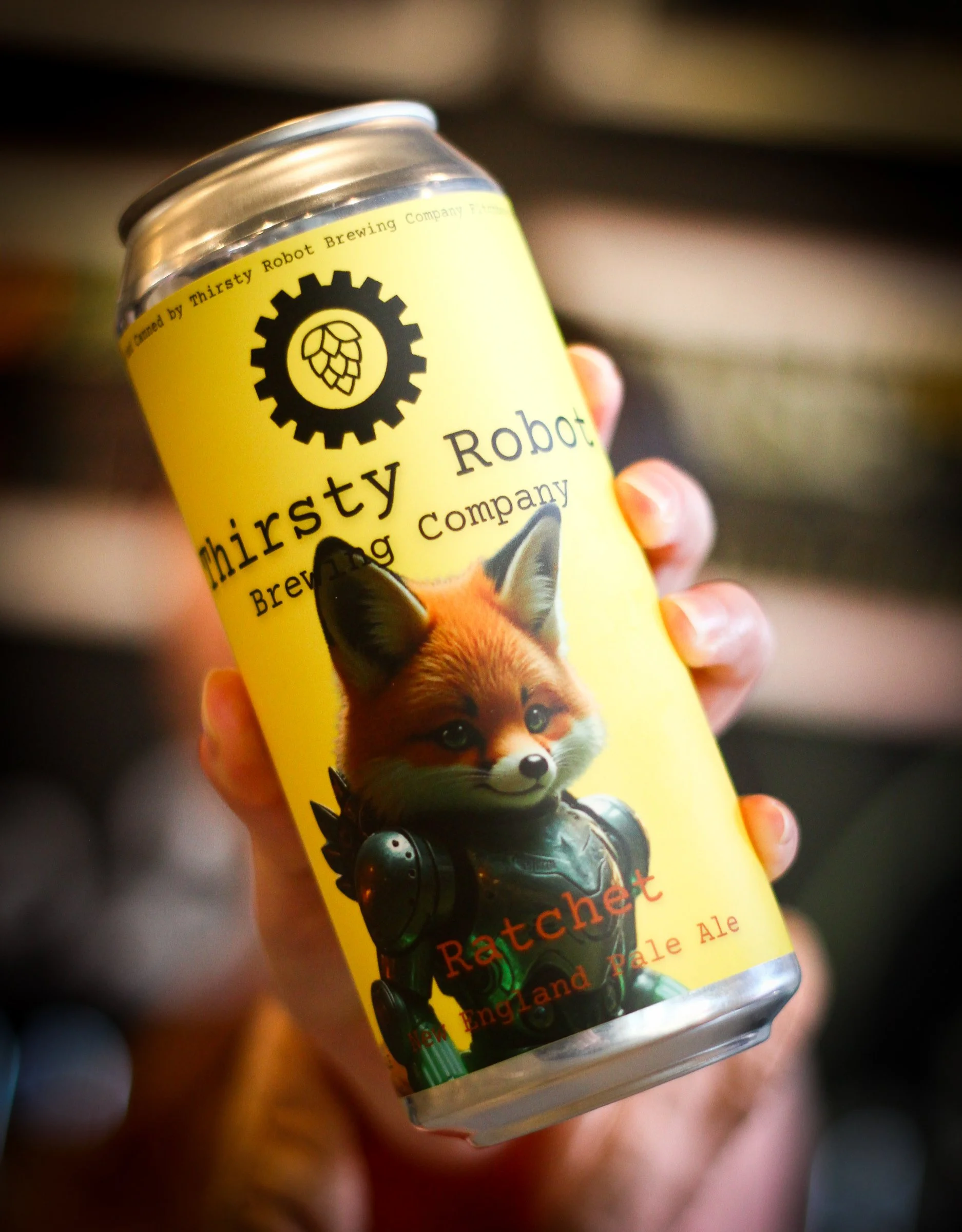 A hand holding a yellow can of Thirsty Robot Brewing Company Ratchet Pale Ale with a fox illustration and a robotic design.
