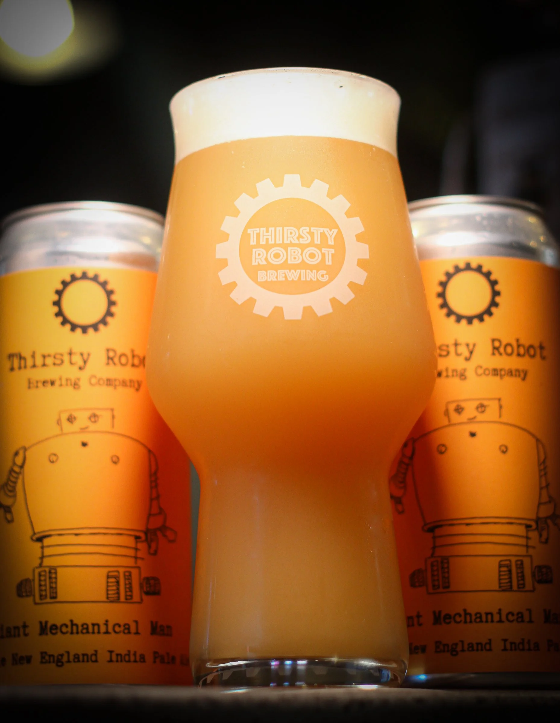 A glass of pale beer with a foamy head in front of two cans of Thirsty Robot Brewing Company beer, featuring a robot drawing on yellow labels.