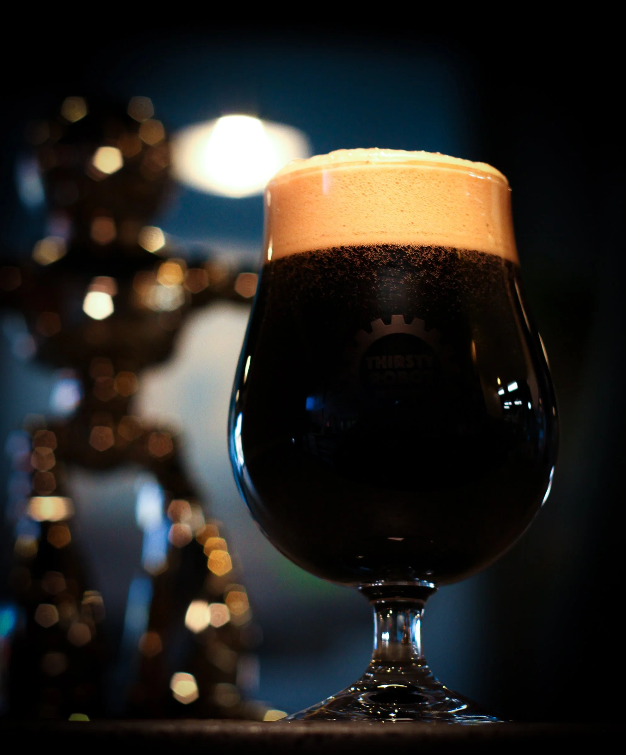 A dark stout beer with a creamy tan head in a tulip glass, with a blurred craft beer tap handle in the background and a bright light behind the glass.