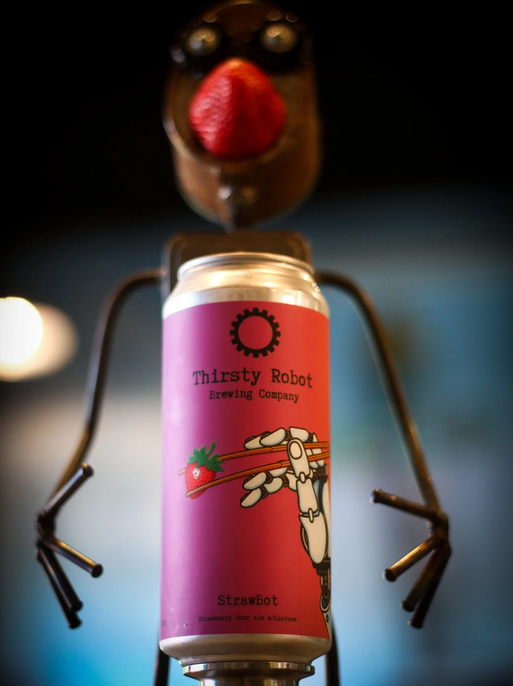 A robot holding a can of Thirsty Robot brewing company Strawberry Sour Ale with a strawberry on top, with a beverage glass in the background.