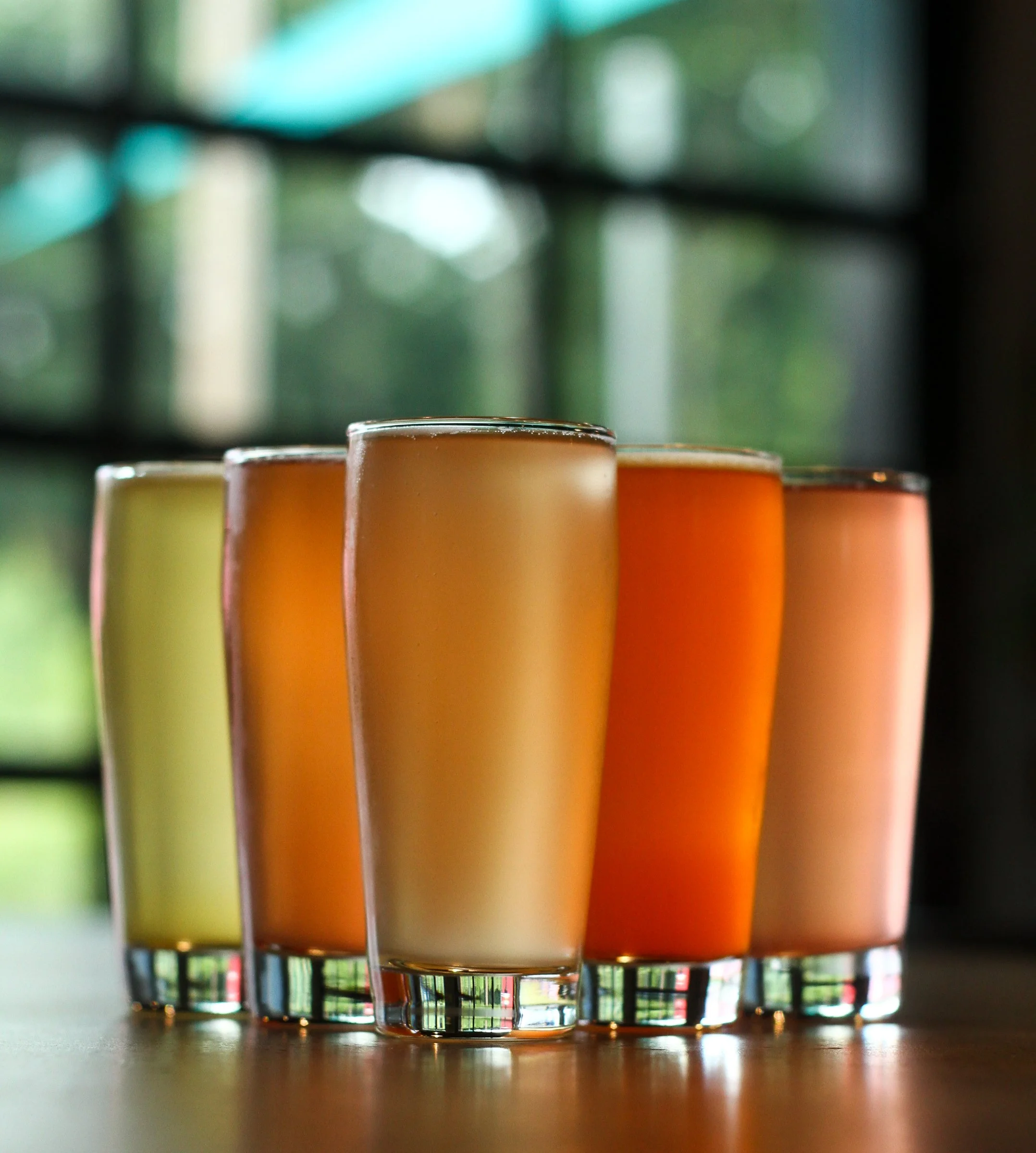 Six glasses of smoothies in different colors, arranged in a row on a wooden surface, with a blurred window in the background.
