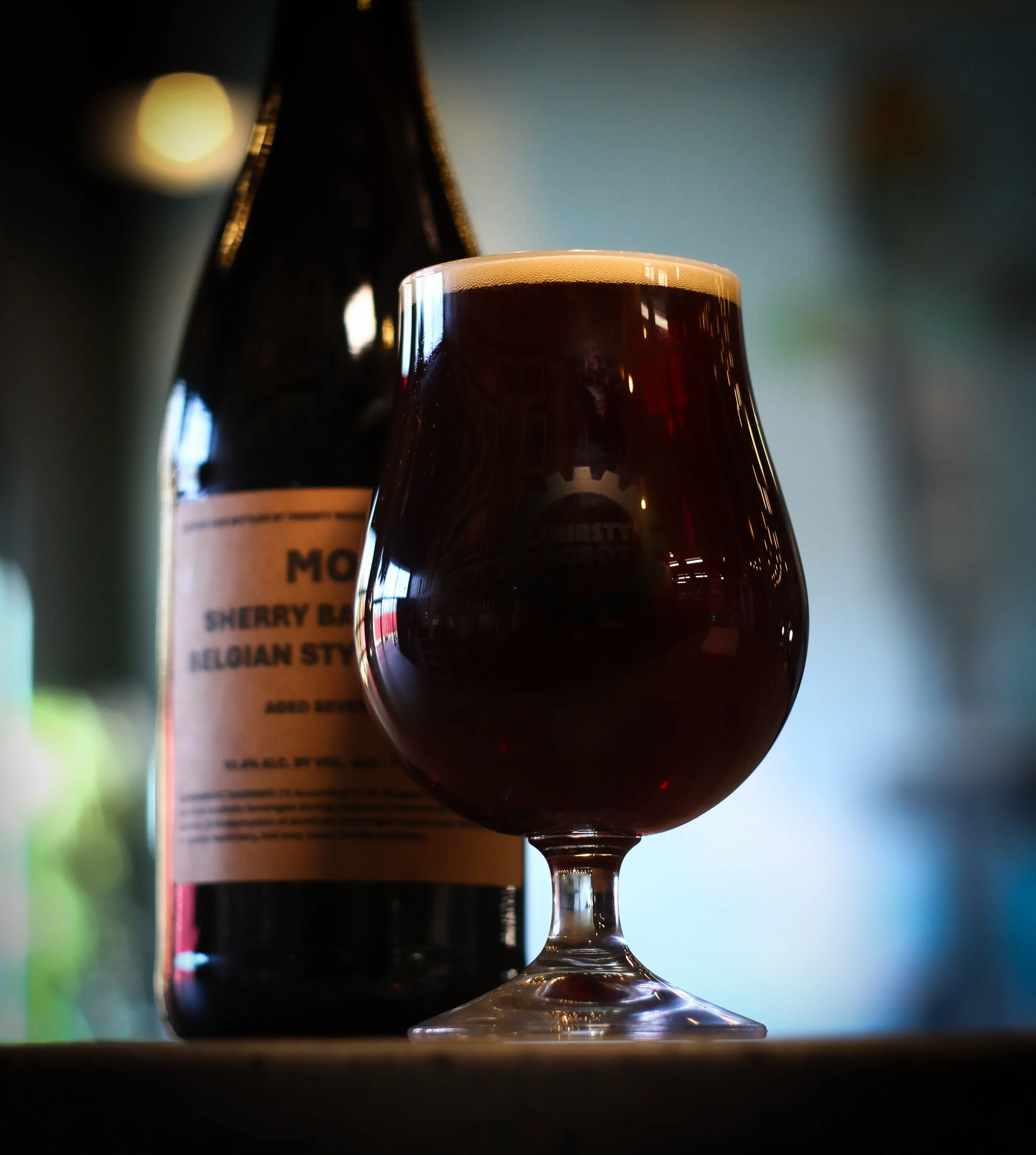 A filled glass of dark red beer with a frothy head, alongside a dark beer bottle labeled 'Cherry Belgian Style' in the background.