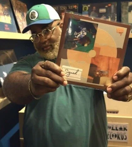 Man wearing glasses and a colorful hat holding a signed artwork in a store, with shelves and signs in the background.