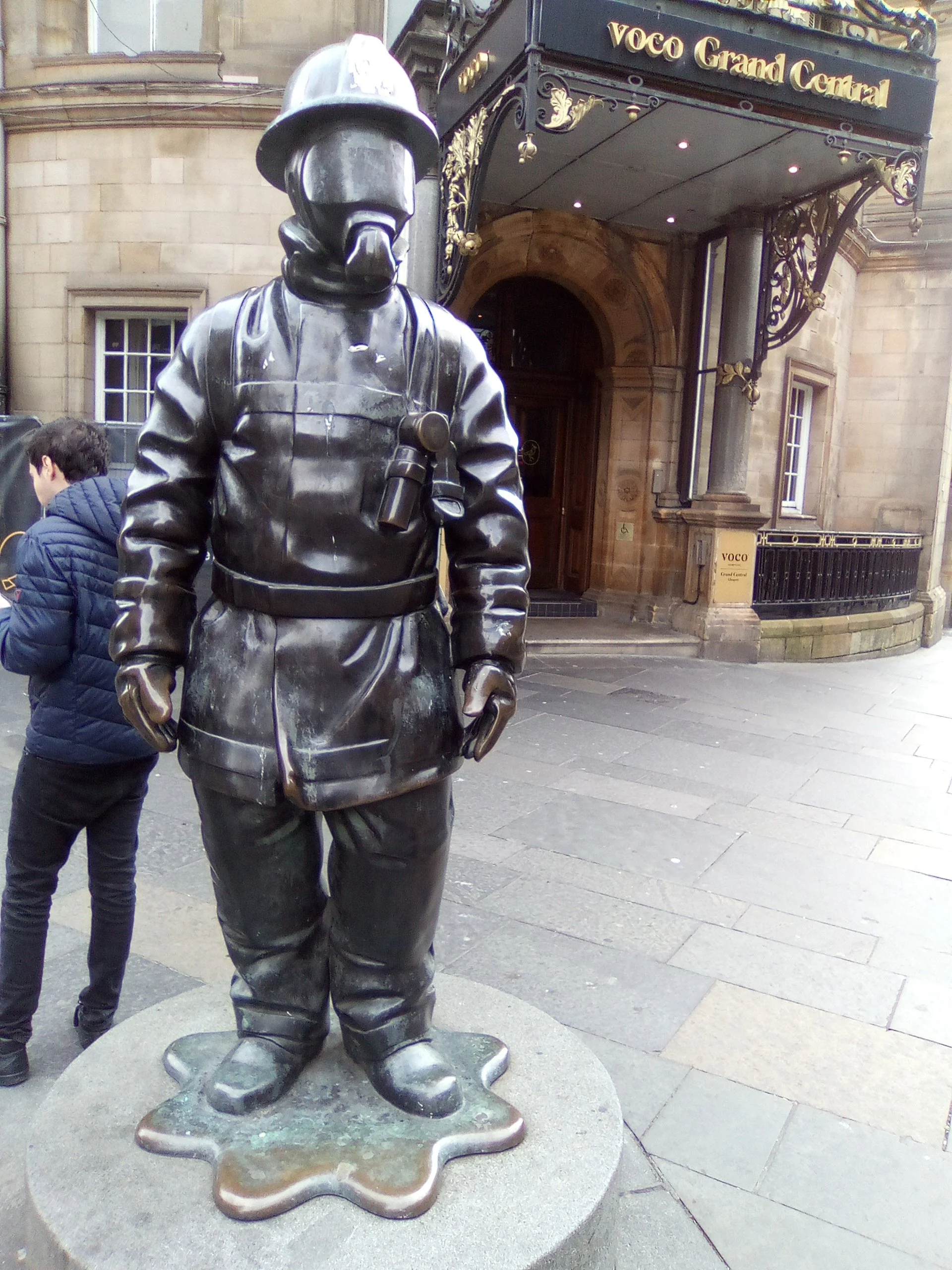 Bronze statue of a Scottish police officer in uniform, standing on a circular base outside a building named VOCO Grand Central.