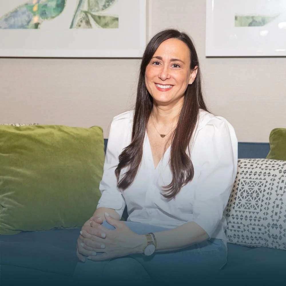 A woman with long brown hair, wearing a white blouse, sitting on a green couch in a room with light-colored walls and framed artwork, smiling at the camera.