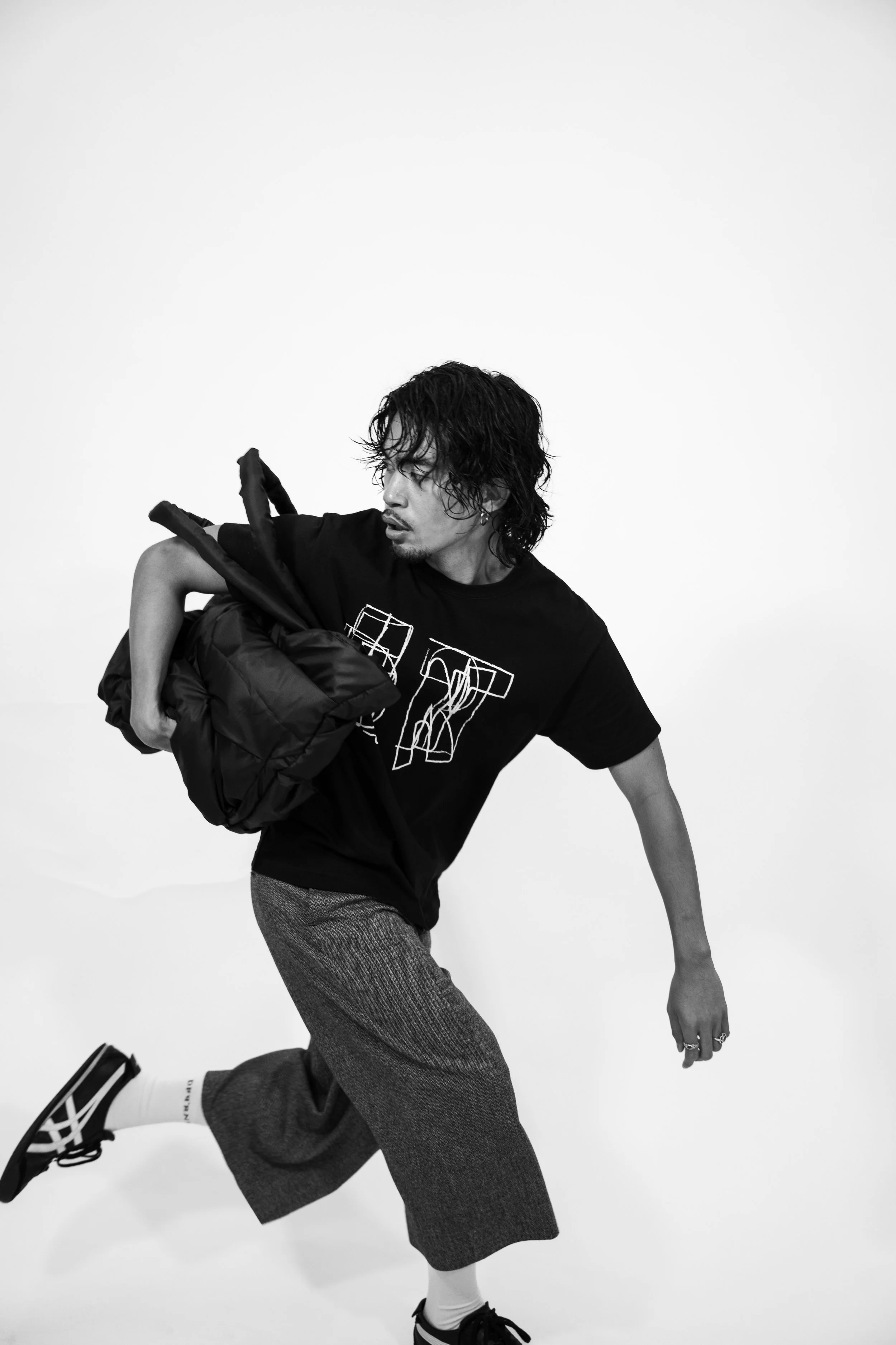 A young man with curly, shoulder-length hair, wearing a black t-shirt, gray wide-leg pants, white socks, and black sneakers, is in motion, carrying a large black bag over his shoulder, against a plain white background.