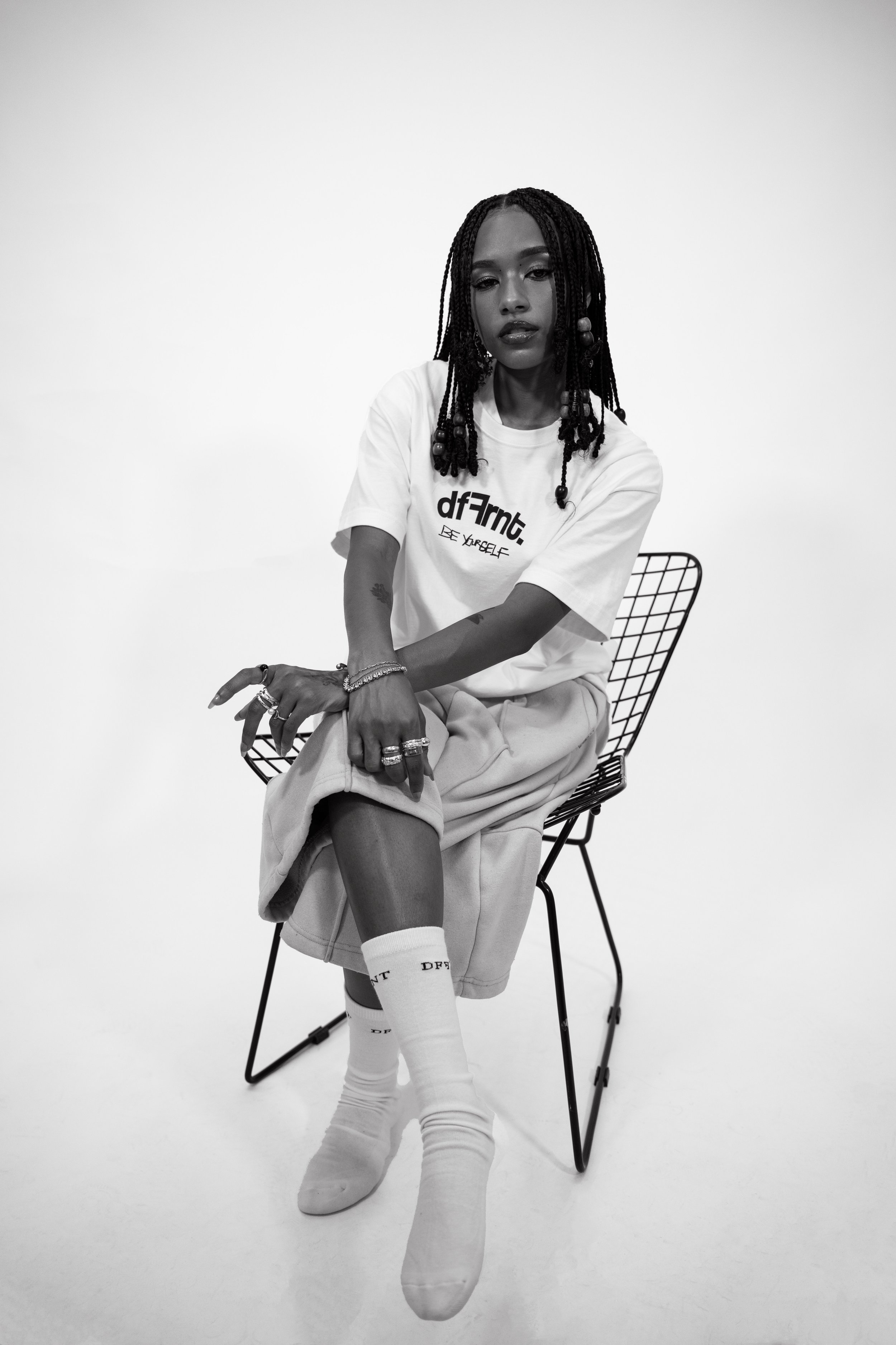 A young woman with braided hair sitting on a wire chair against a plain background. She is wearing a white t-shirt, beige pants, and white socks, with multiple rings and bracelets.