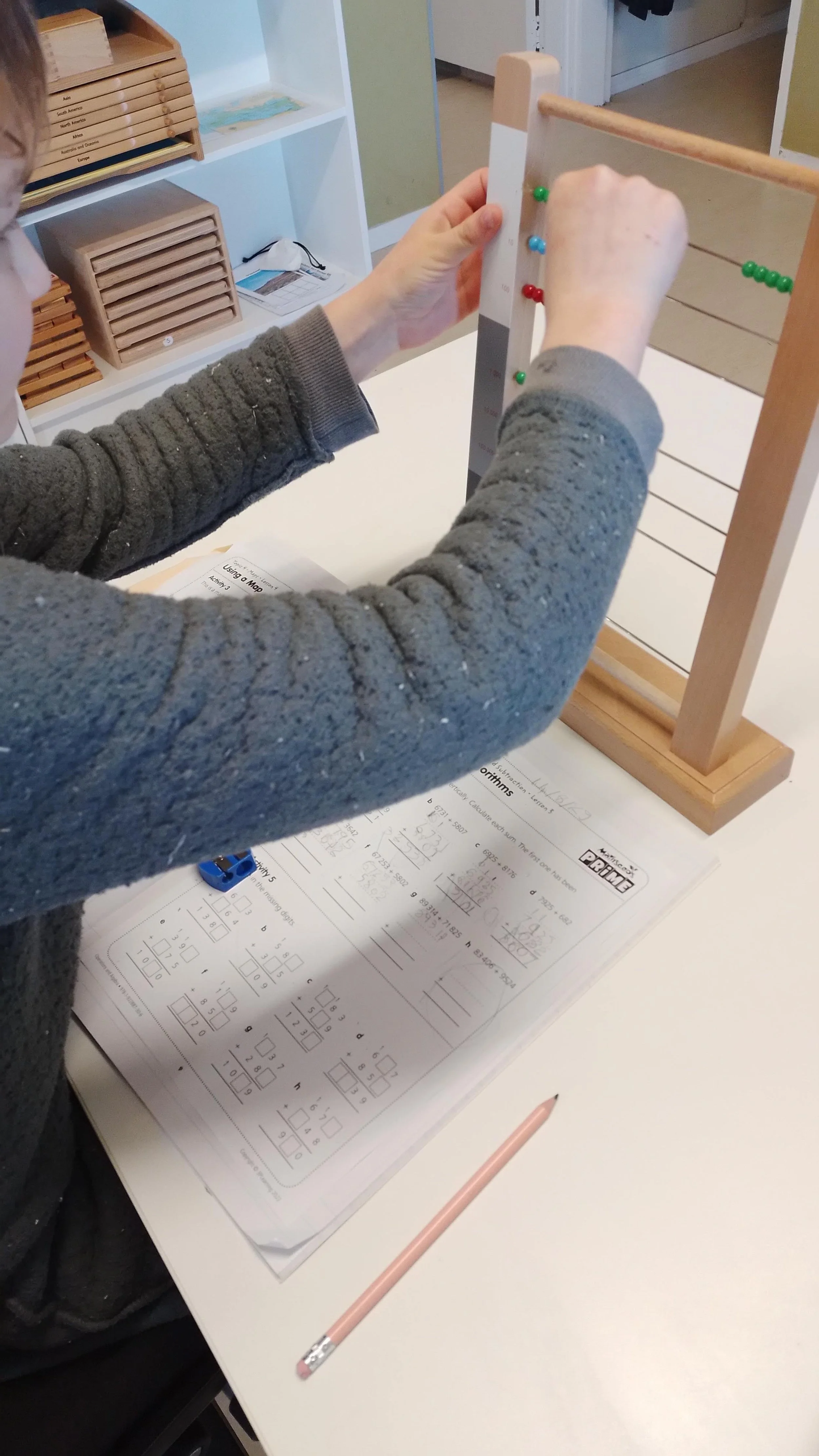 Child working on a math worksheet while using a montessori teaching tool of a learning activity at a white table in a classroom.
