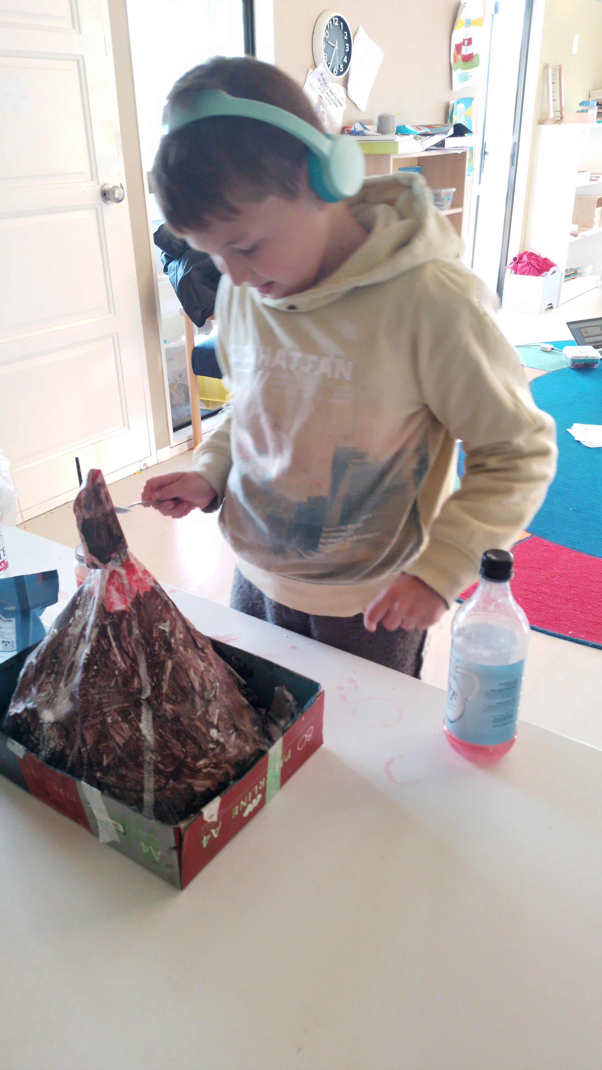 A young boy wearing a cream hoodie and teal headphones standing at a table undertaking science experiment. neurodivergent learning enviornment.