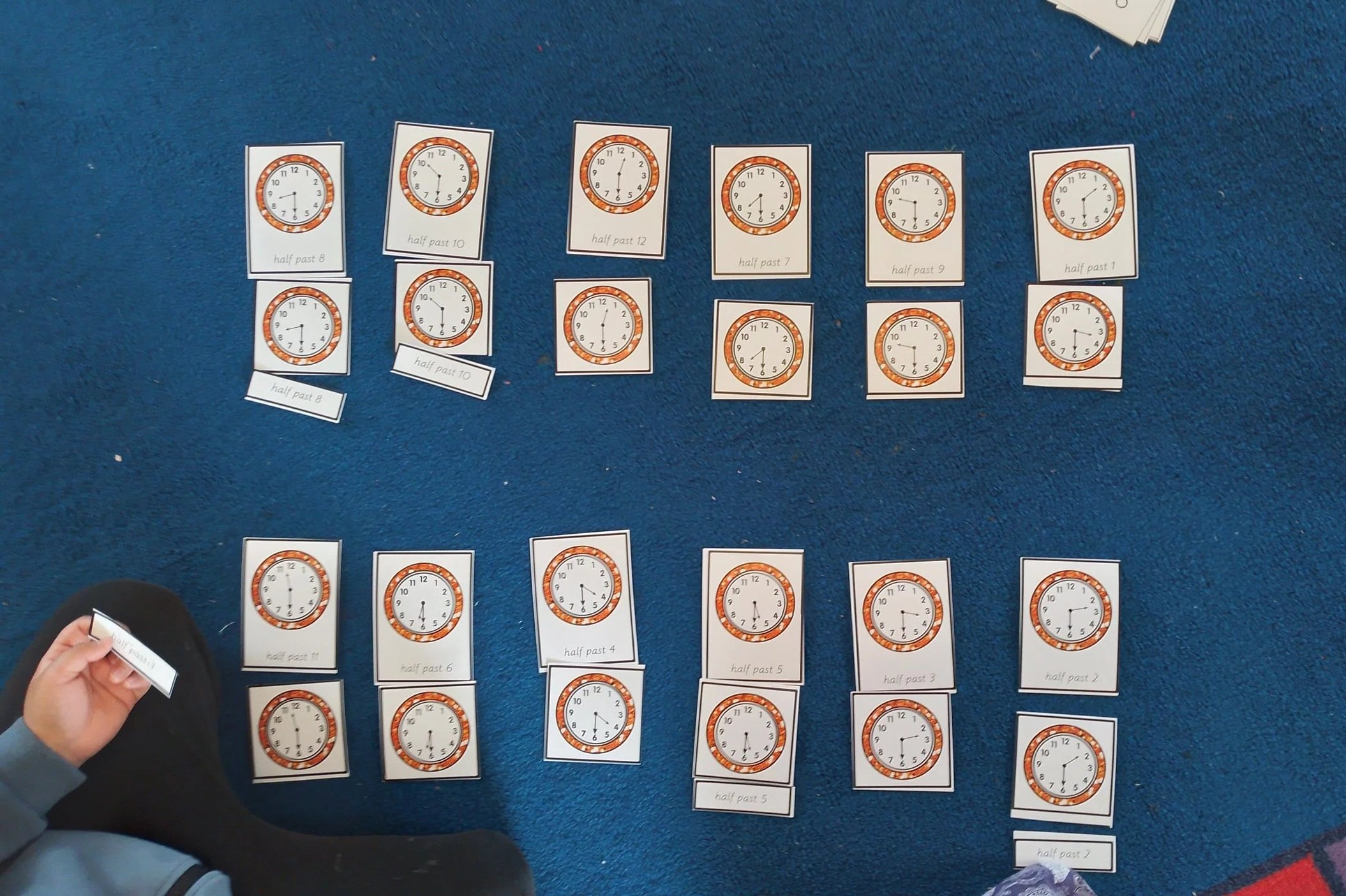 Collection of clock cards laid out on a blue carpet, with some cards labeled with times like 'half past 8', 'half past 10', and 'half past 12'. A child is holding one card labeled 'half past 11'.