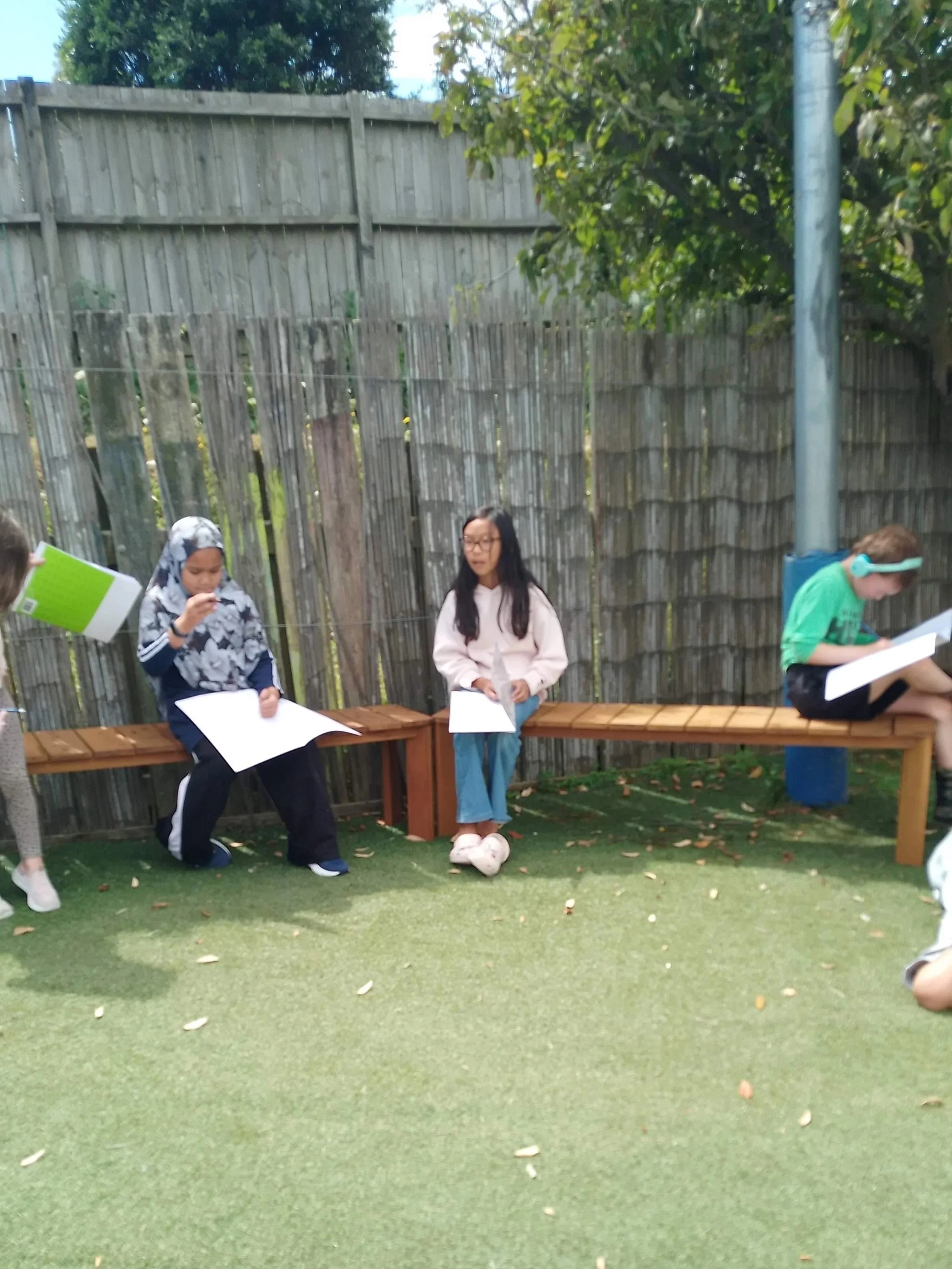Kids sitting on benches outside, reading and talking, in a yard with a wooden fence and trees.