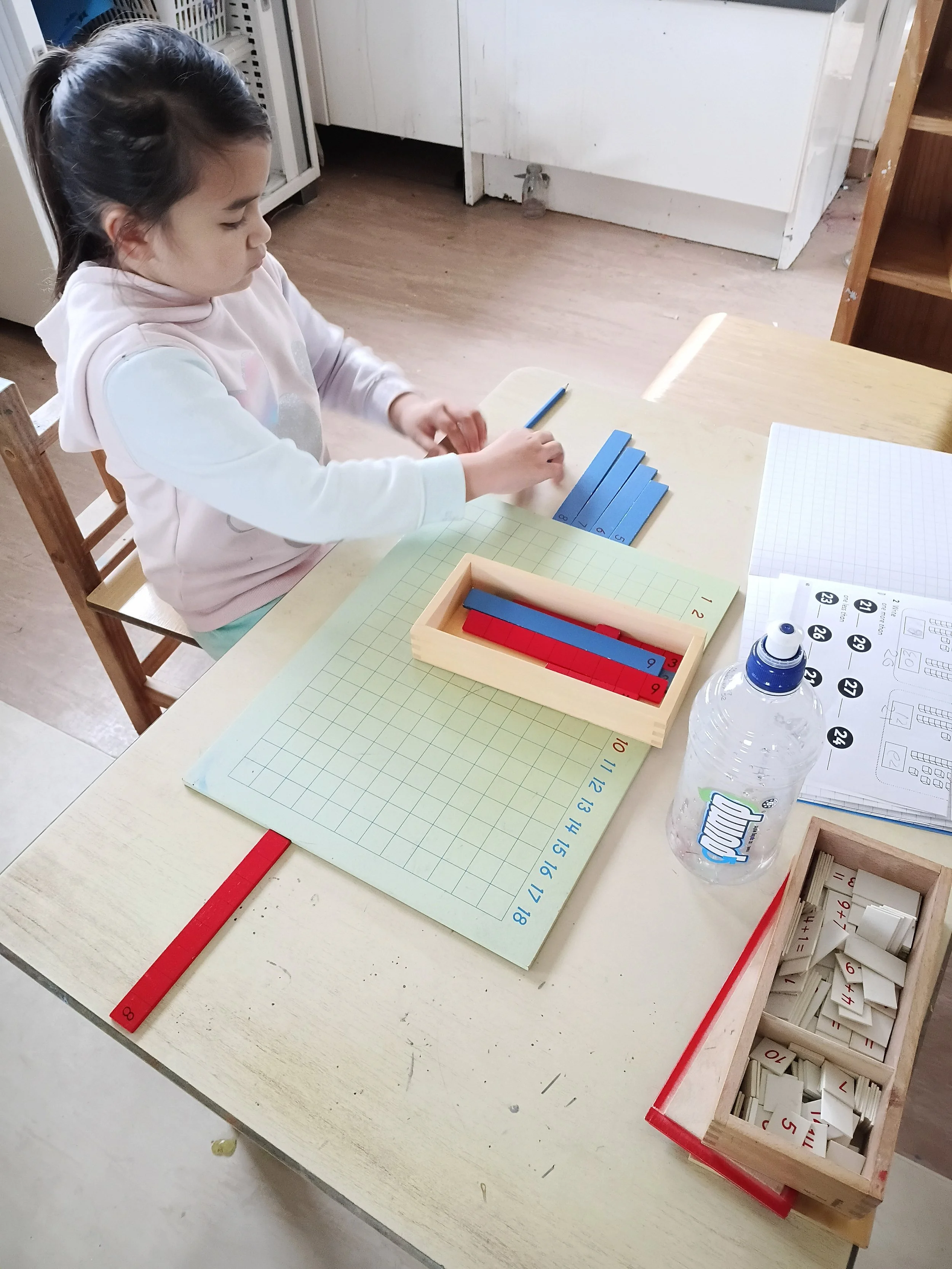 Girl sitting at a table with educational materials, including number tiles, a water bottle, and a worksheet.